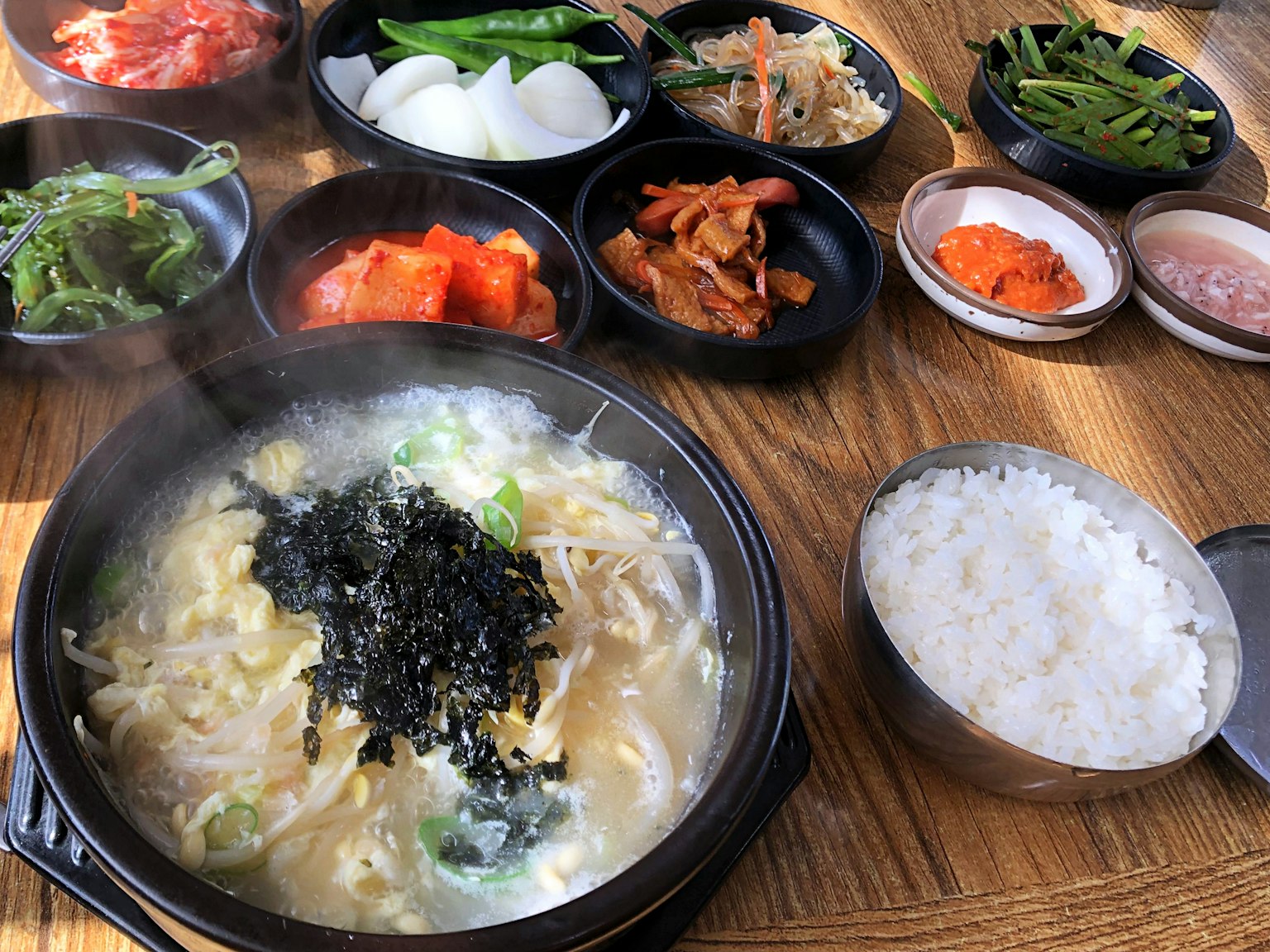 Eine dampfende Schüssel Suppe mit Reis umgeben von verschiedenen koreanischen Beilagen