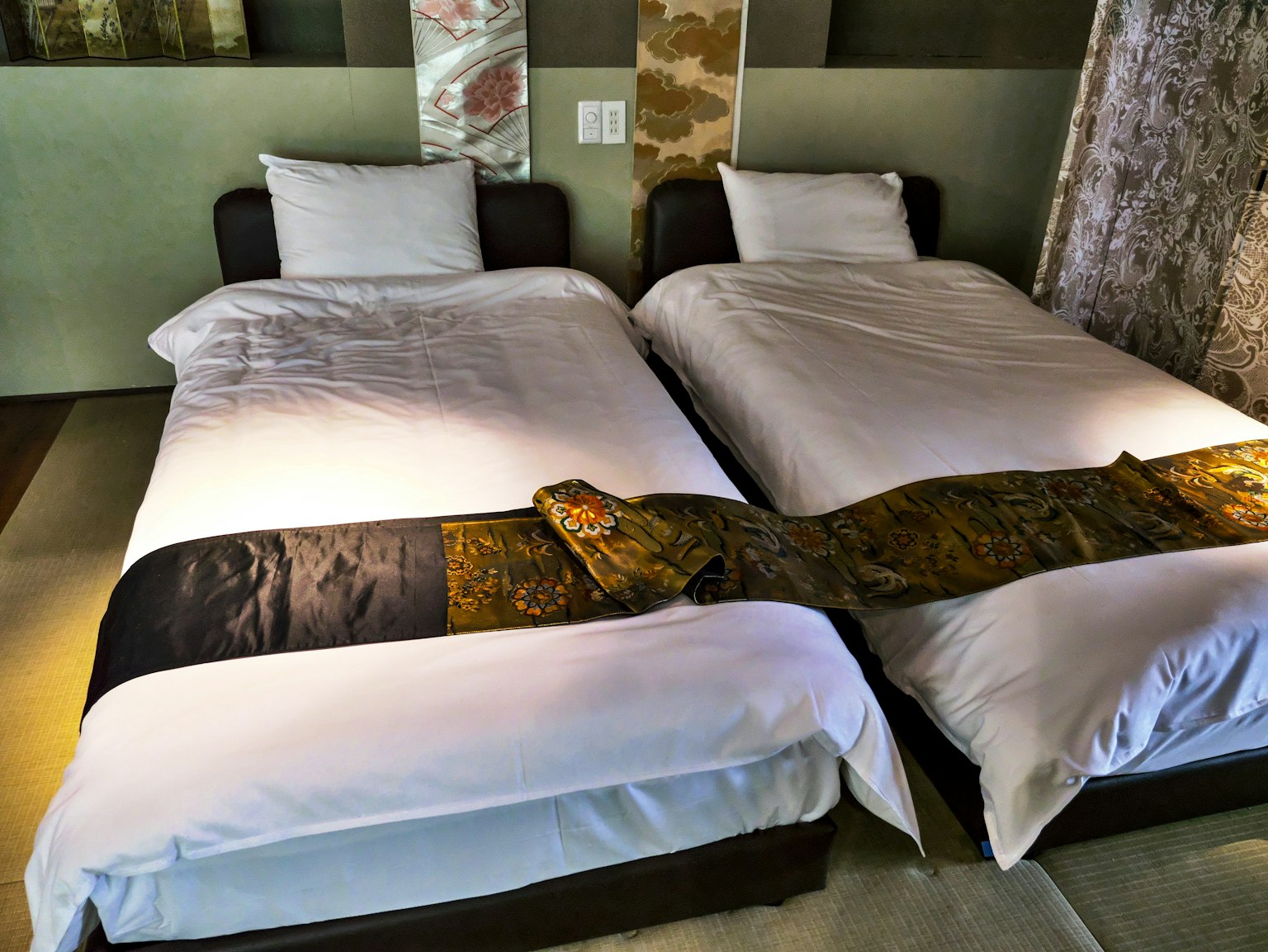 Dua tempat tidur tunggal di dalam ruangan modern dengan seprai putih dan kain dekoratif berwarna-warni di atasnya