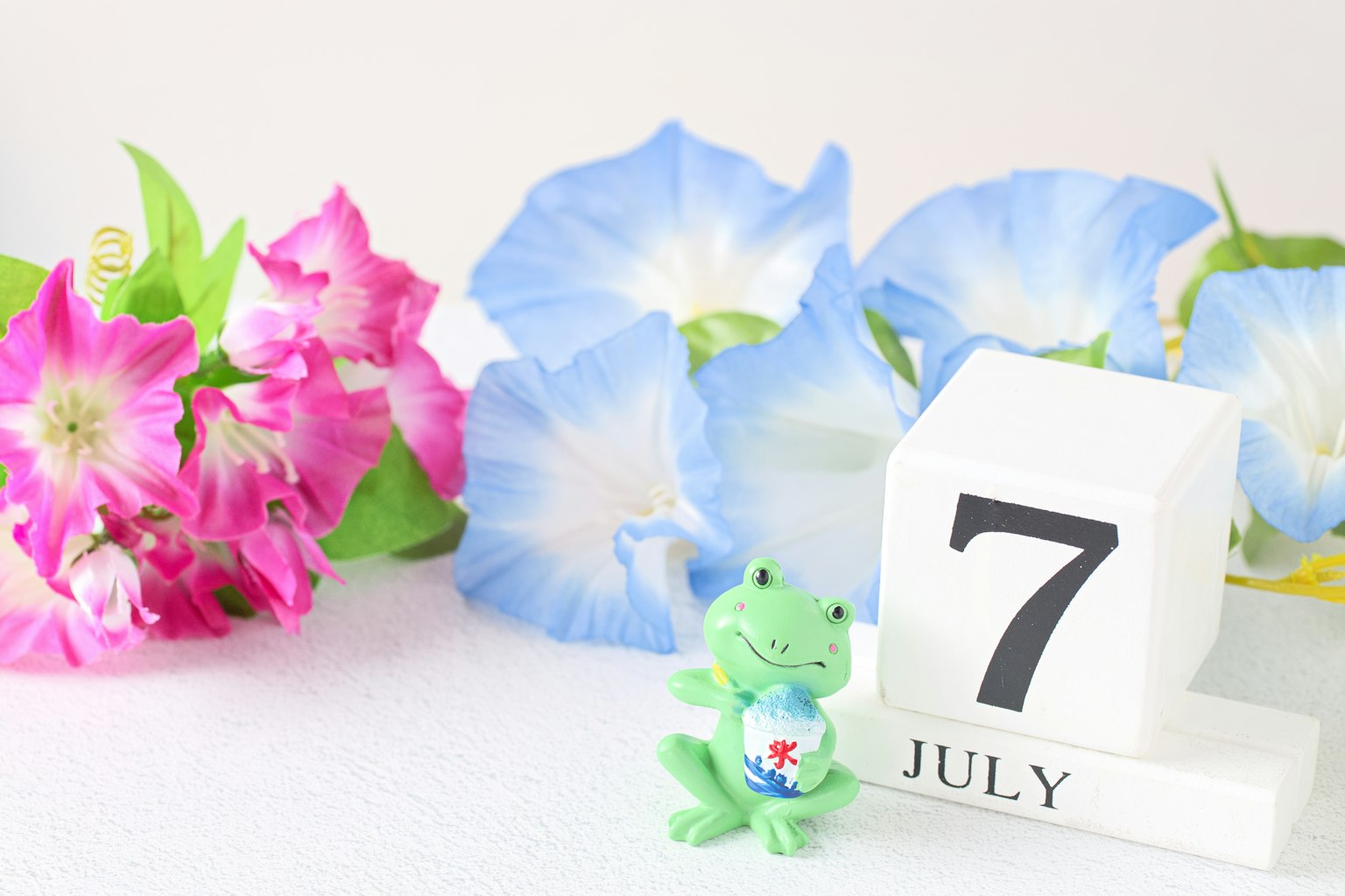顯示7月7日的日曆，周圍環繞著藍色牽牛花和粉色花朵，以及一個青蛙擺飾