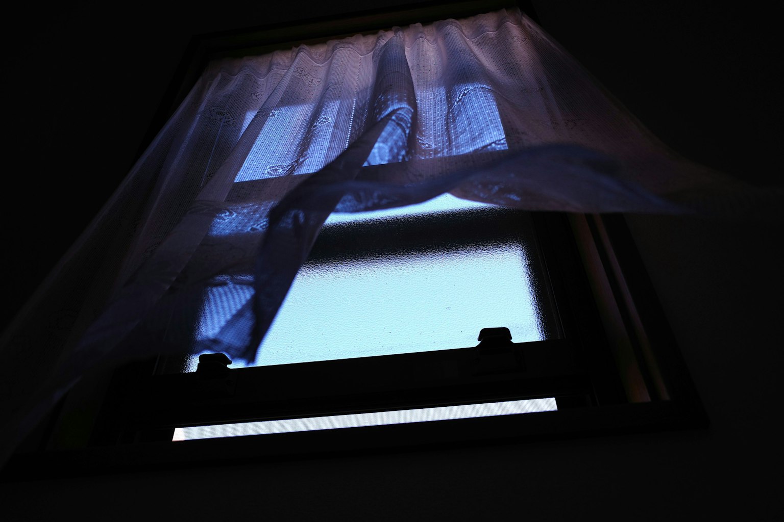 Cửa sổ trong một căn phòng tối, hé lộ bầu trời xanh và rèm trắng đung đưa trong gió