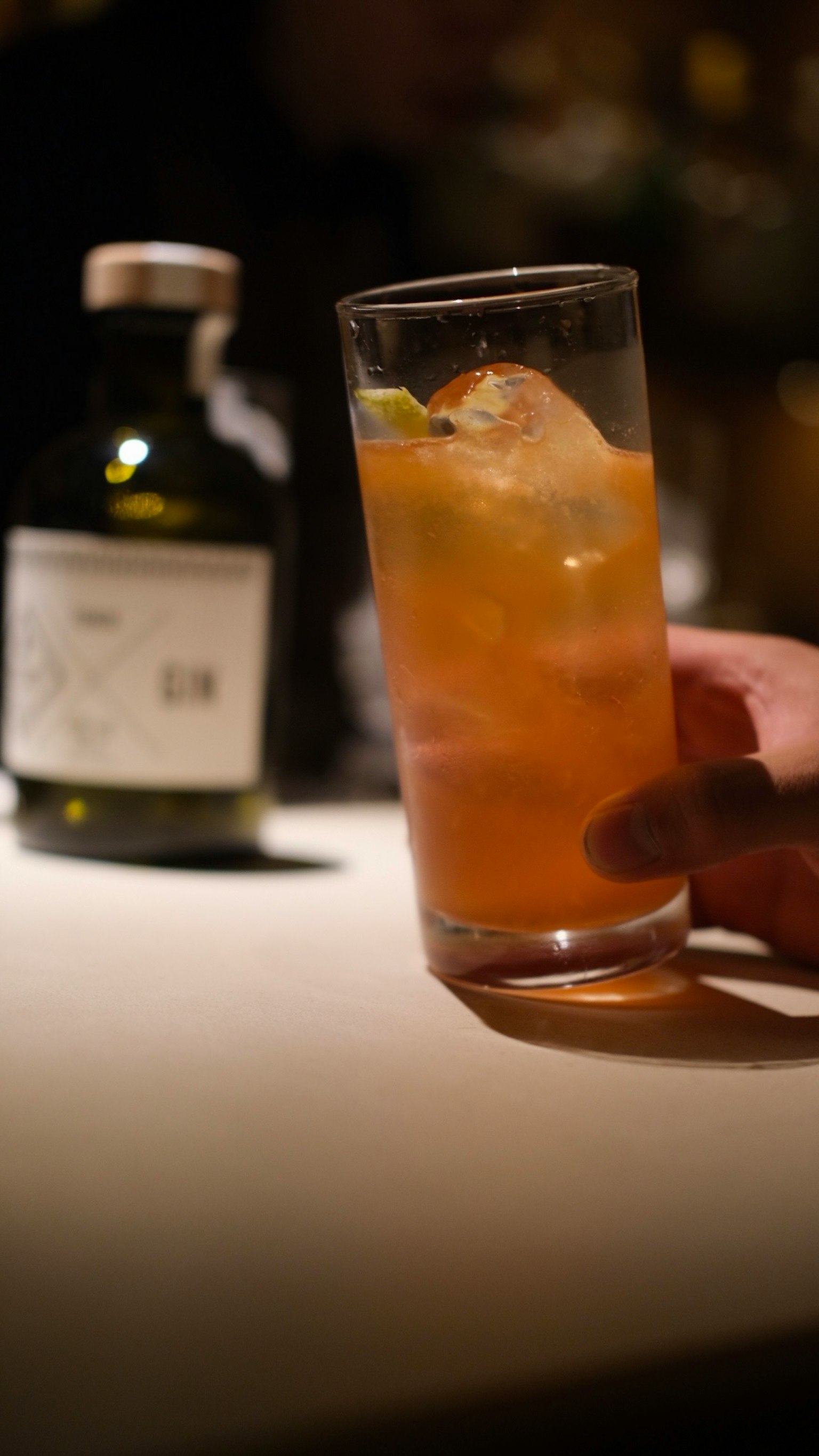 Una mano che tiene un bicchiere di cocktail con ghiaccio e una bottiglia sullo sfondo