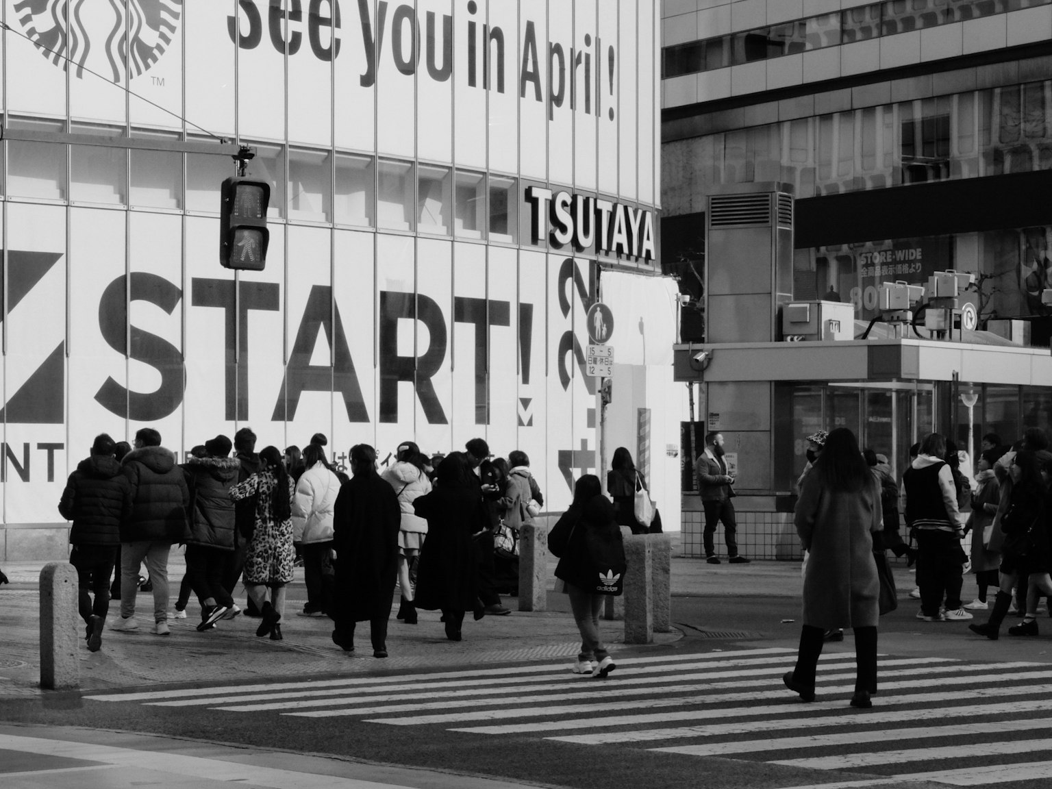 Des personnes traversant la rue dans un paysage urbain noir et blanc avec le panneau TSUTAYA et le texte START