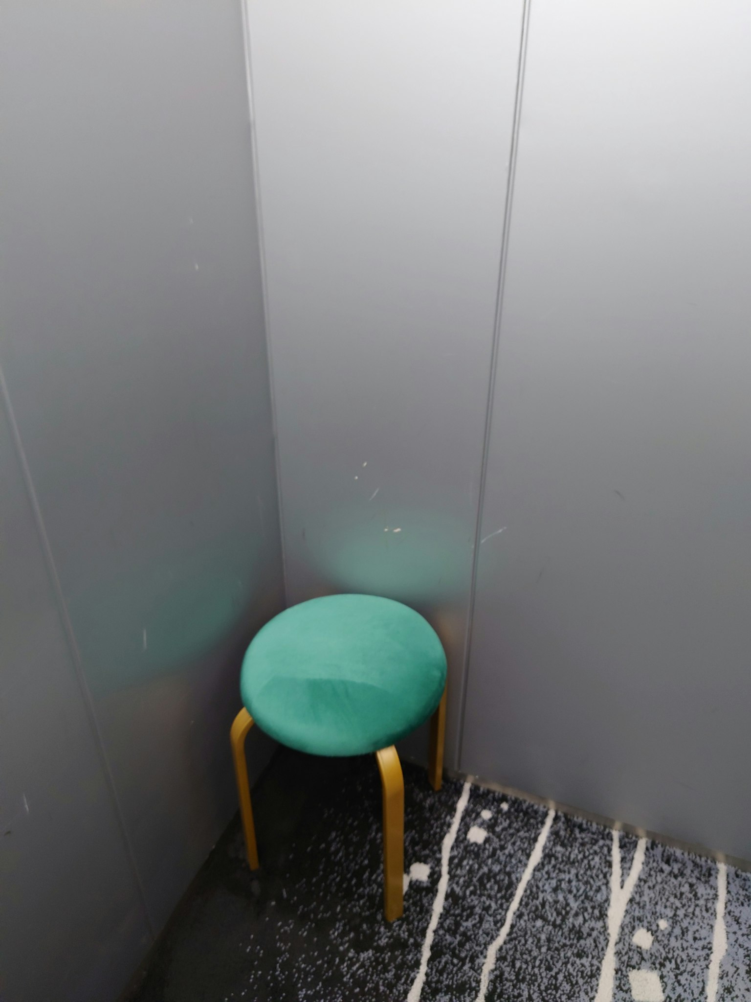 A green cushioned stool in a gray-walled corner