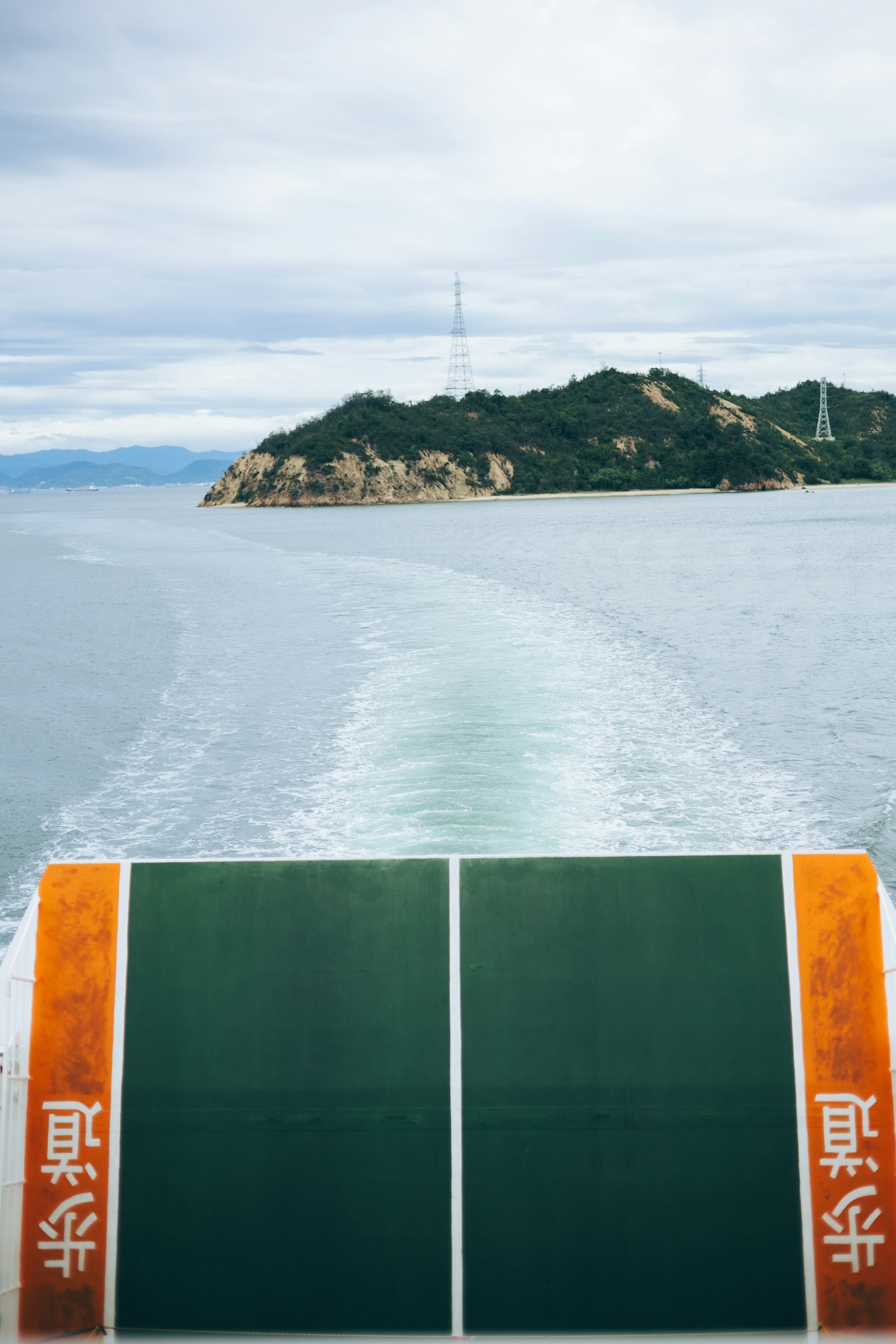 View from the back of a boat showing the sea and an island Boat deck is visible with mountains and a communication tower on the island