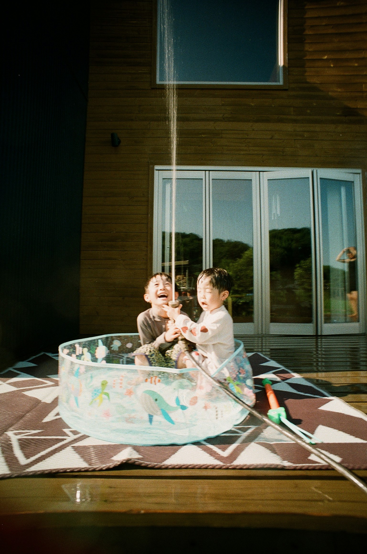 เด็ก ๆ กำลังเล่นในสระน้ำขนาดเล็กที่เป่าลมพร้อมบ้านไม้ในพื้นหลัง