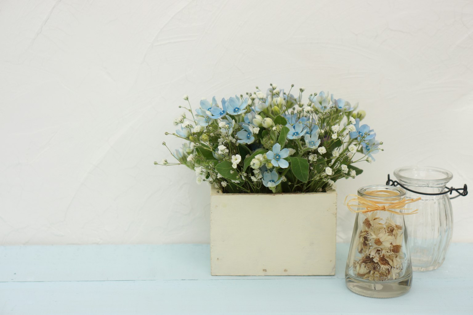 Una maceta con flores azules y flores blancas junto a frascos de vidrio sobre una superficie azul clara