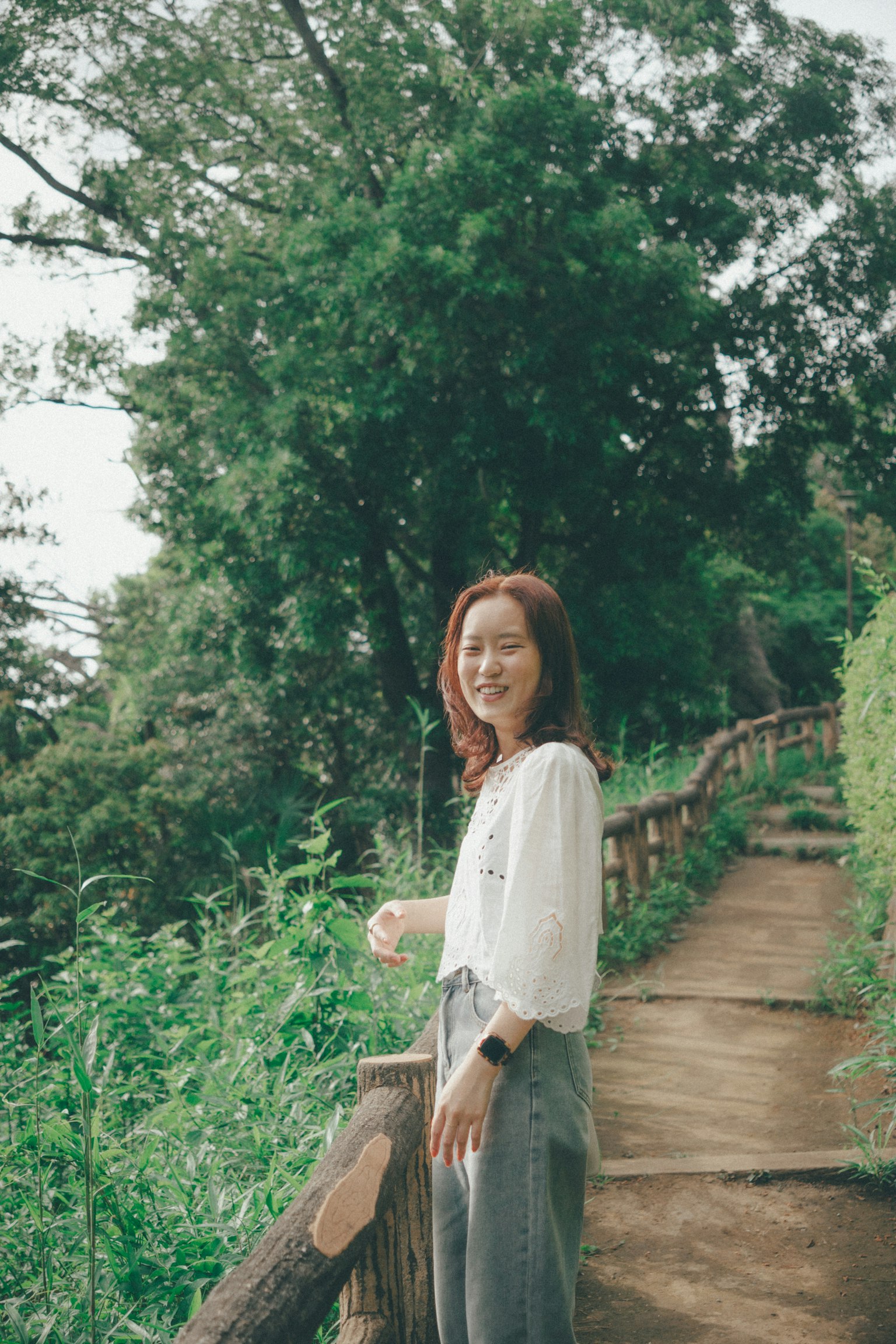 Người phụ nữ mỉm cười trên con đường được bao quanh bởi cây cối mặc áo trắng và váy xám