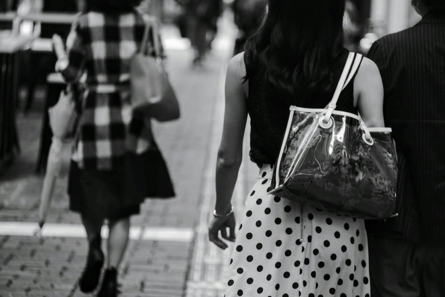 Seorang wanita mengenakan gaun polkadot berjalan di jalan dengan sosok lain di latar belakang