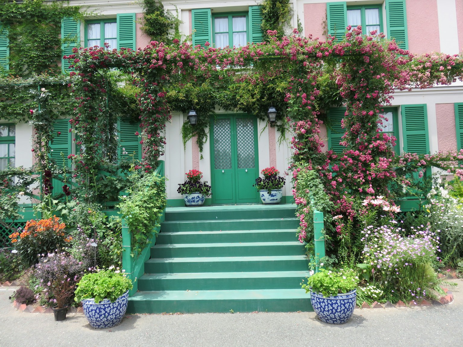 Maison rose avec portes et fenêtres vertes ornée de plantes et de fleurs luxuriantes