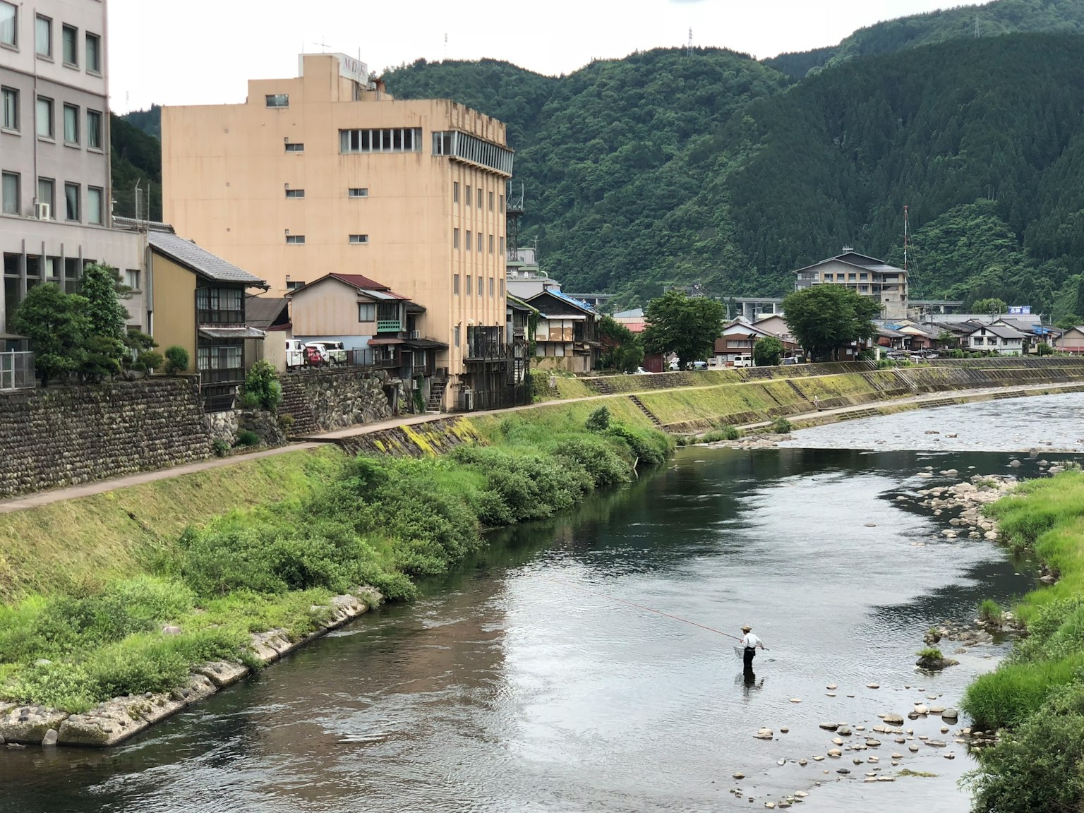 A person fishing in a river with mountains and buildings in the background