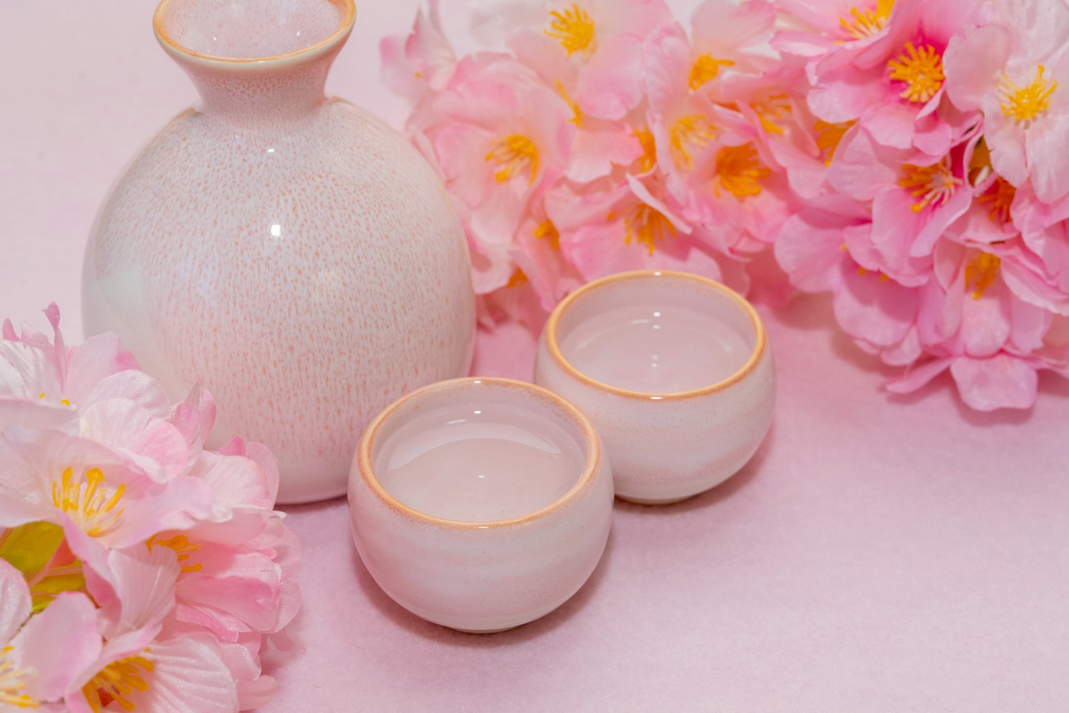 Bình sake bằng gốm trắng và hai chén nhỏ được bao quanh bởi hoa hồng nhạt