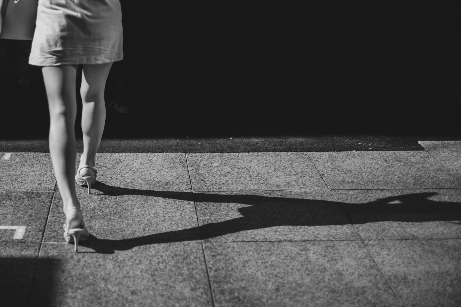 Photo en noir et blanc d'une femme marchant sur le trottoir ses jambes projetant une ombre au sol