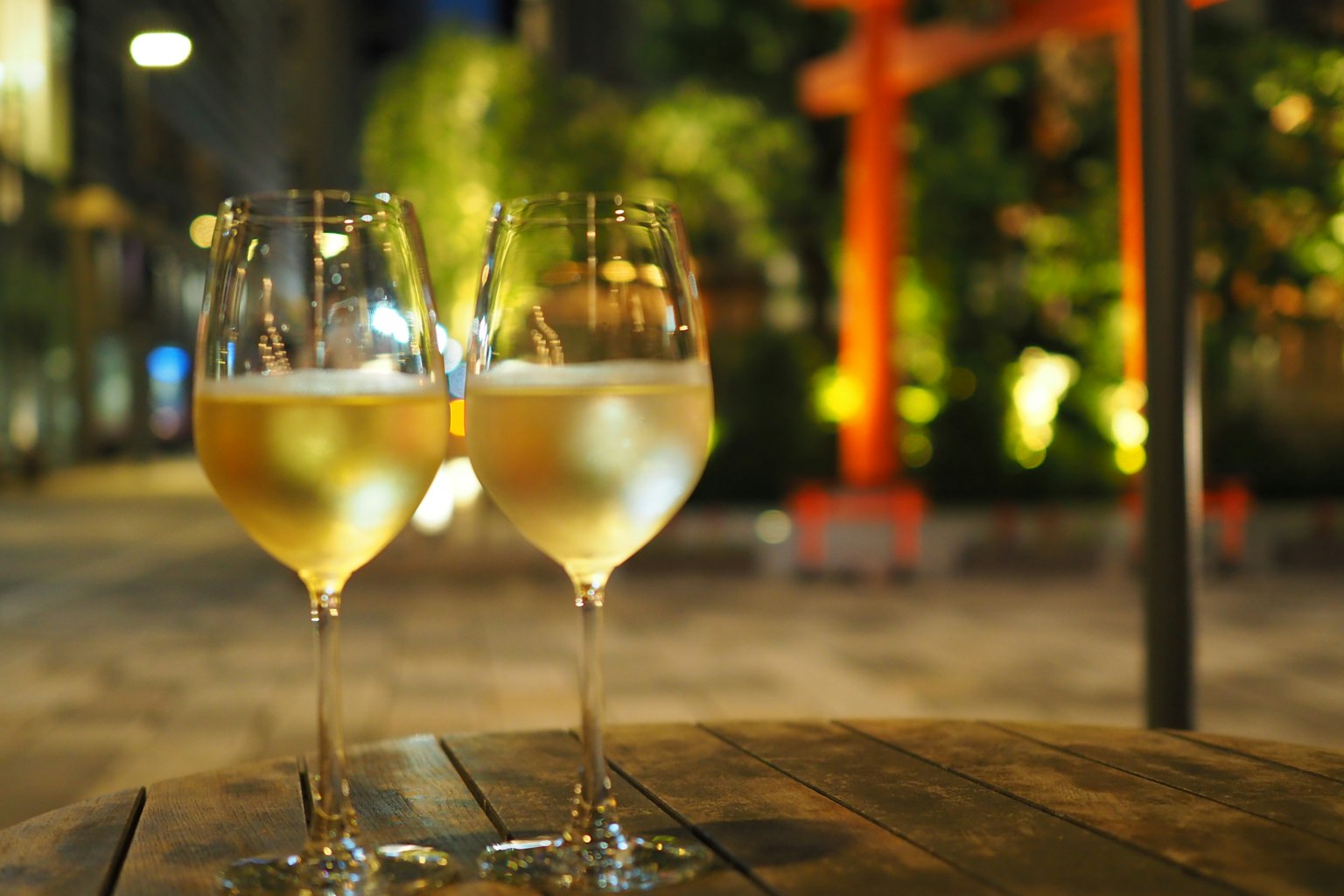 两只装有白葡萄酒的酒杯放在木桌上,背景是夜晚的红色鸟居和绿色植物