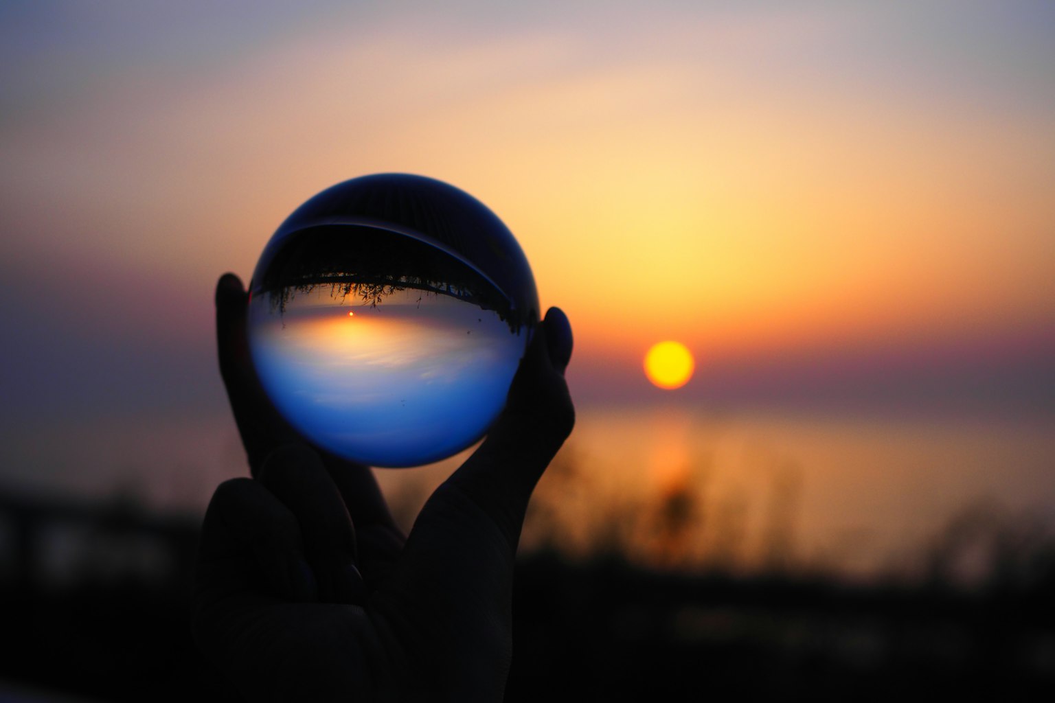 Eine Hand hält eine Glaskugel, die einen Sonnenuntergang über dem Ozean reflektiert