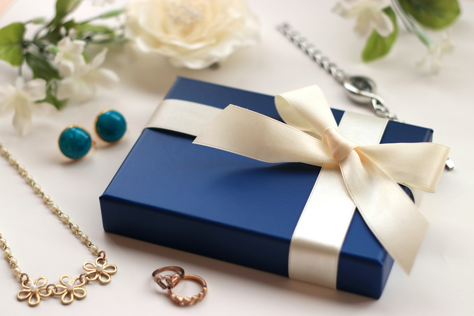 Una scatola regalo blu con un nastro bianco circondata da gioielli e fiori