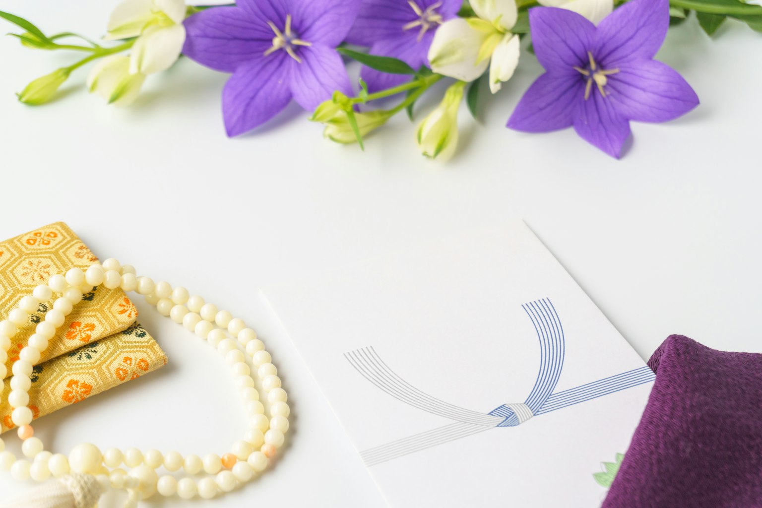 Un arrangiamento di fiori viola e bianchi con una collana di perle e oggetti dorati accanto a una busta decorata con un nastro