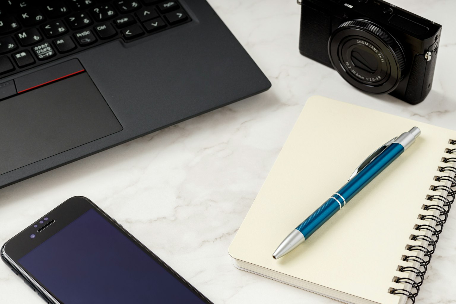 Una macchina fotografica nera accanto a un laptop, uno smartphone, un taccuino e una penna blu