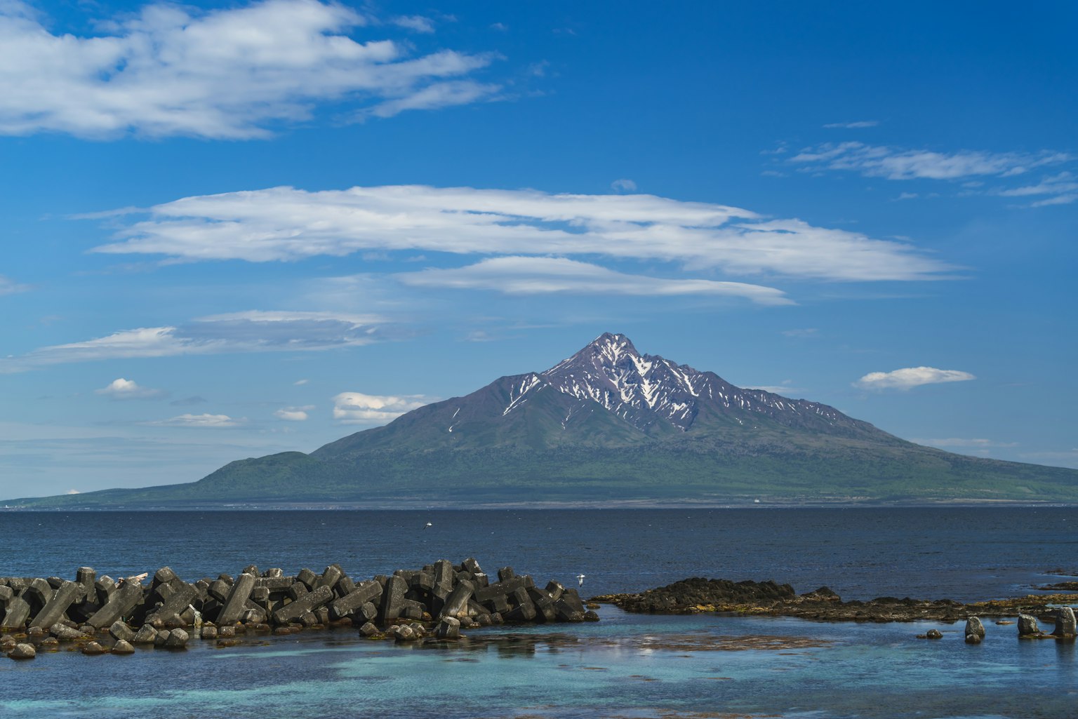 Snow-capped mountain rising under a blue sky with clouds Green slopes and blue sea in the foreground