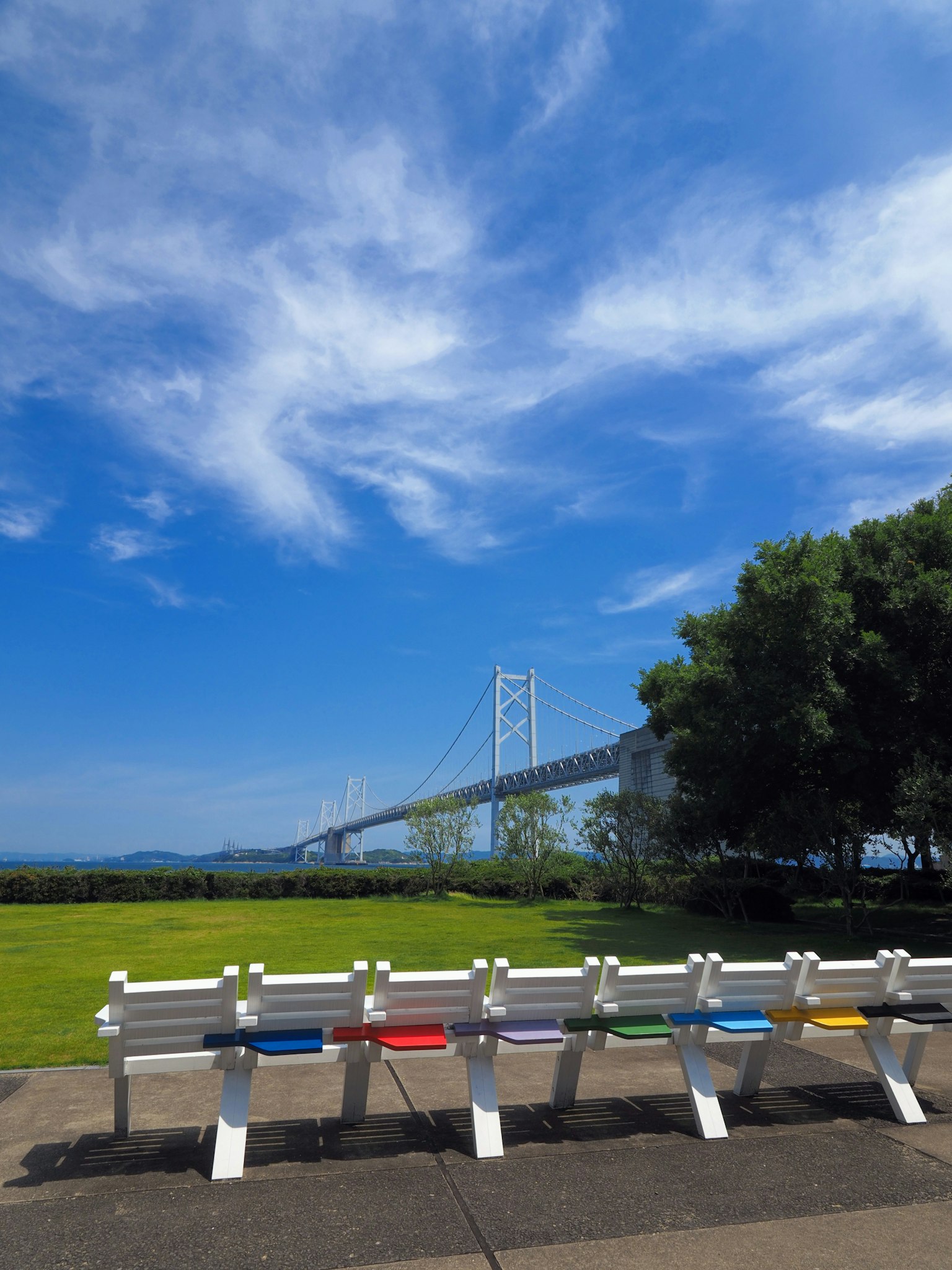 Bunte Bänke unter einem blauen Himmel mit weißen Wolken und einer Brücke im Hintergrund
