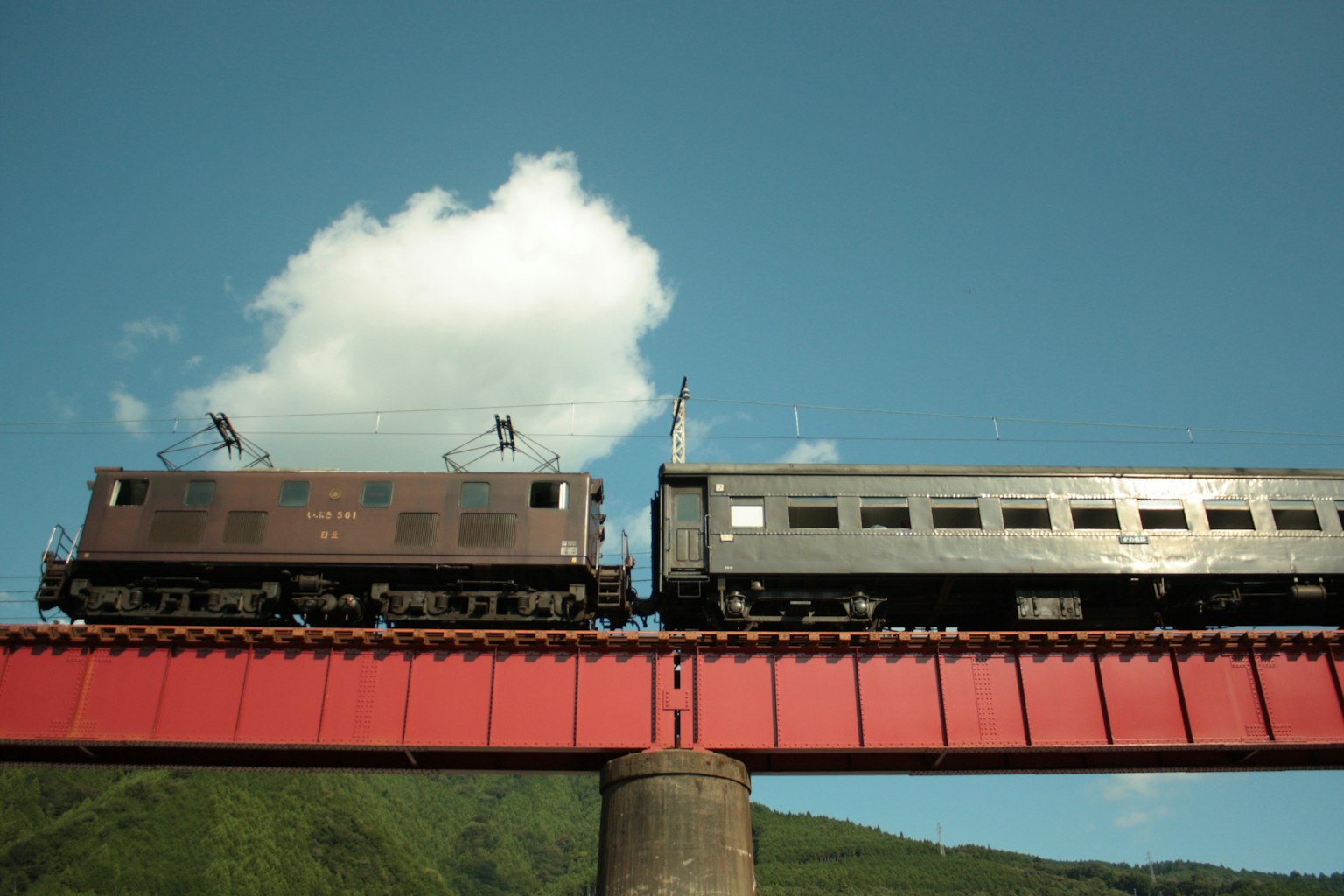 Tren y vagón de pasajeros cruzando un puente rojo bajo un cielo azul con nubes blancas