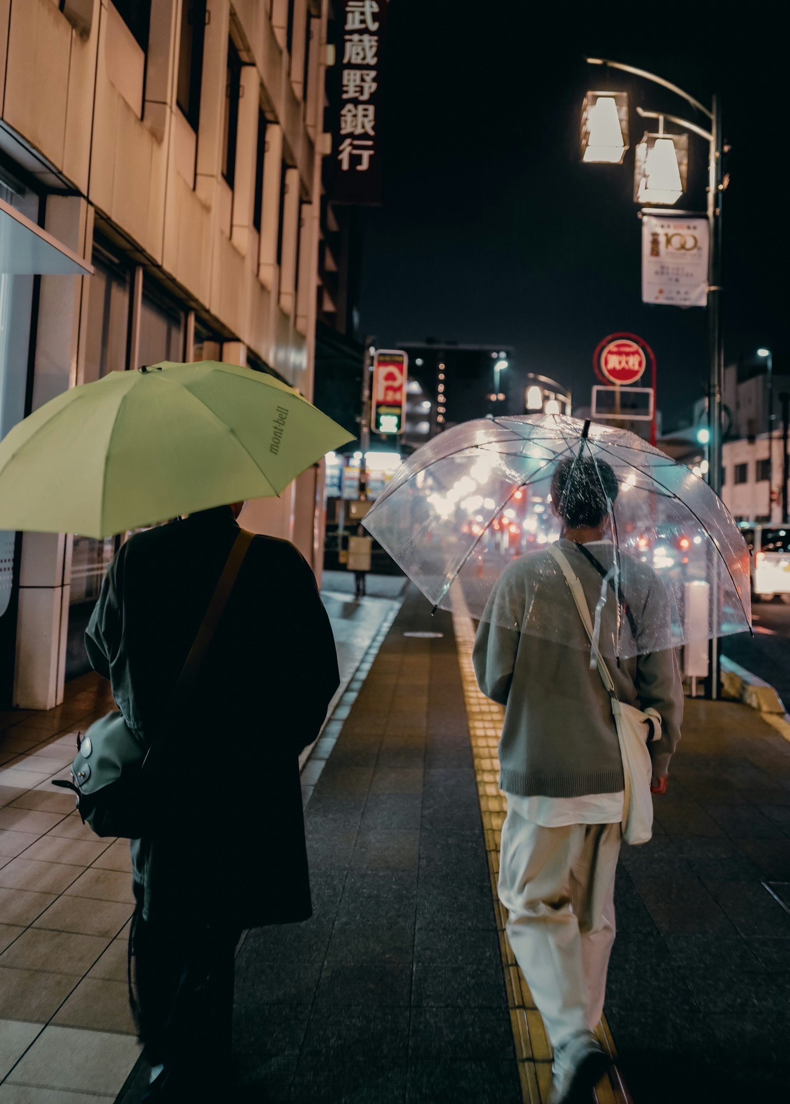 Two people walking in the night city holding a green umbrella and a clear umbrella