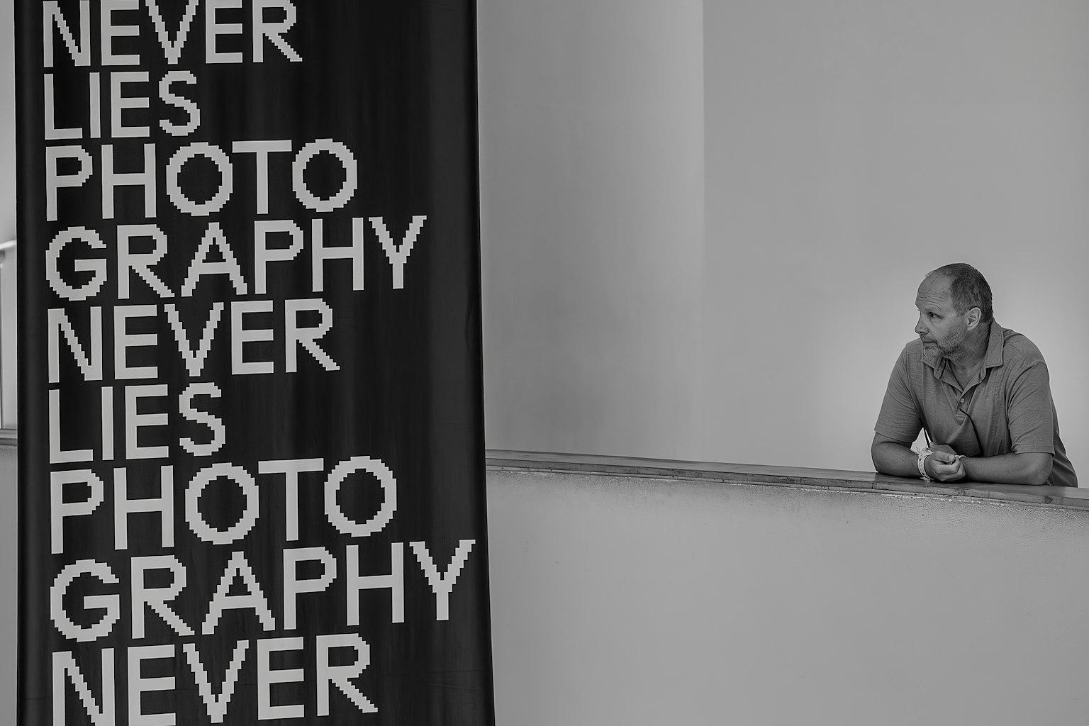 ชายคนหนึ่งยืนอยู่ในพื้นที่ขาวดำข้างข้อความใหญ่ 'NEVER LIES PHOTOGRAPHY'