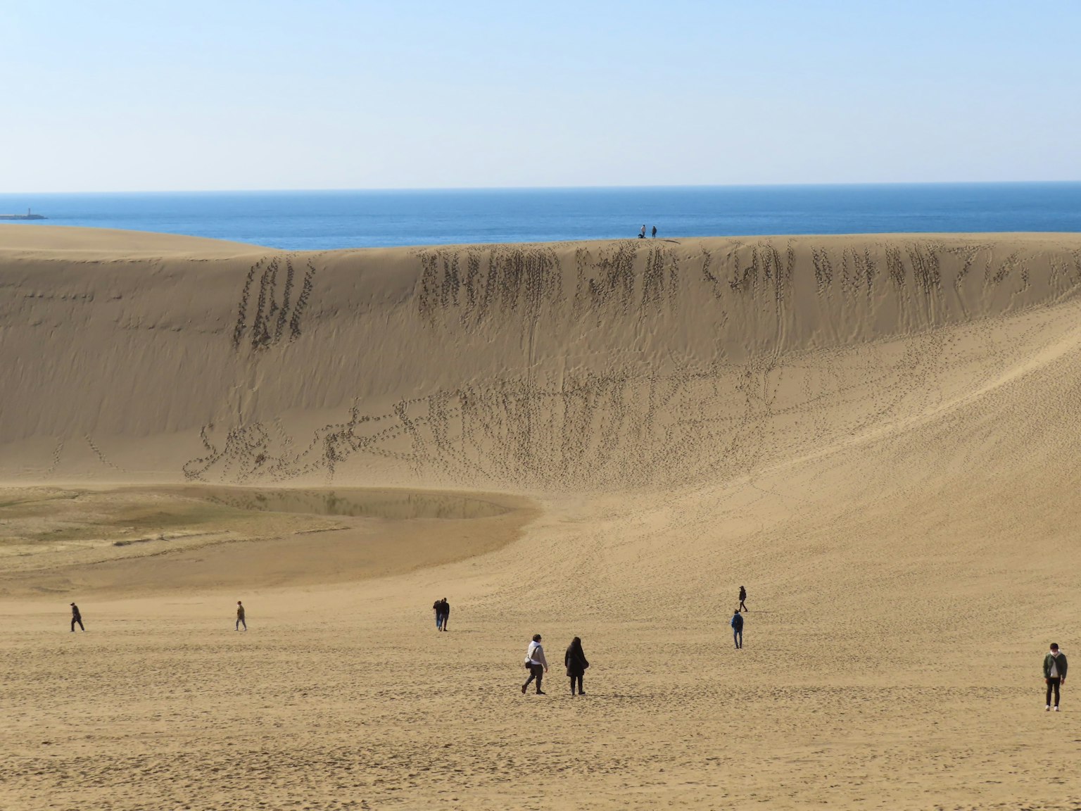Personas caminando sobre vastas dunas de arena con un océano azul al fondo