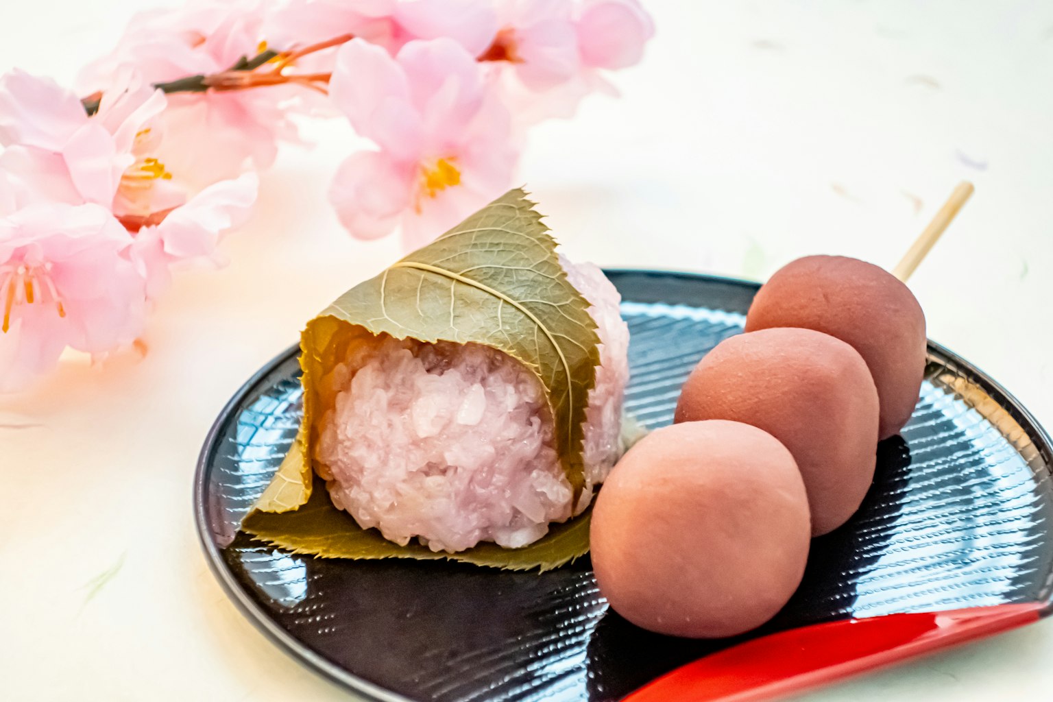 Bola de arroz envuelta en hojas junto a flores de cerezo y tres dango en un plato