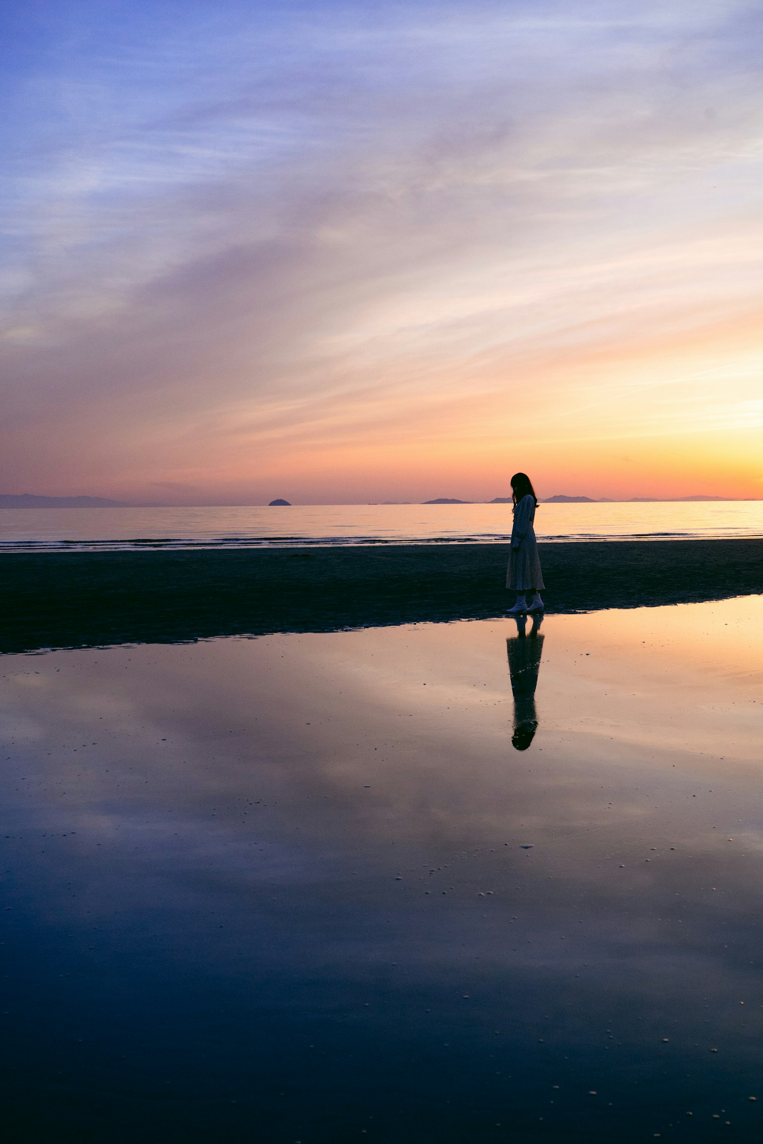 A person standing by the seaside at dusk with their reflection visible in the water