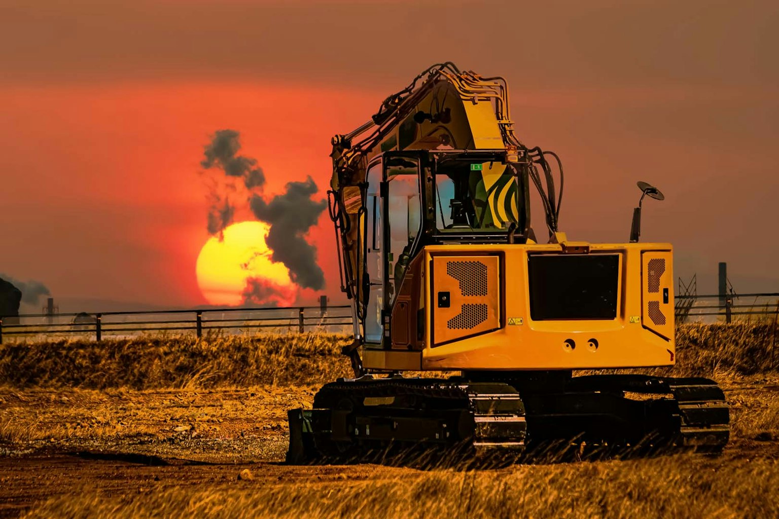 Escavatore giallo contro un tramonto con fumi sullo sfondo