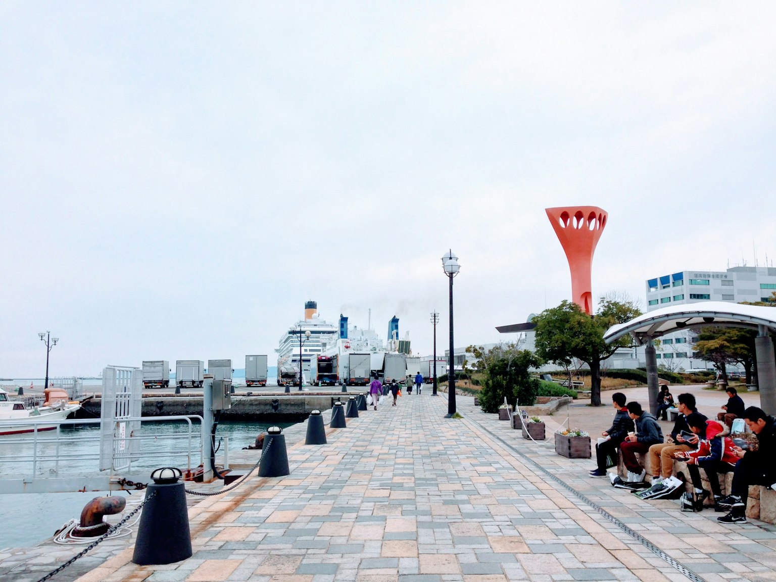 Seaside walkway with people sitting and a port view featuring a red tower and boats