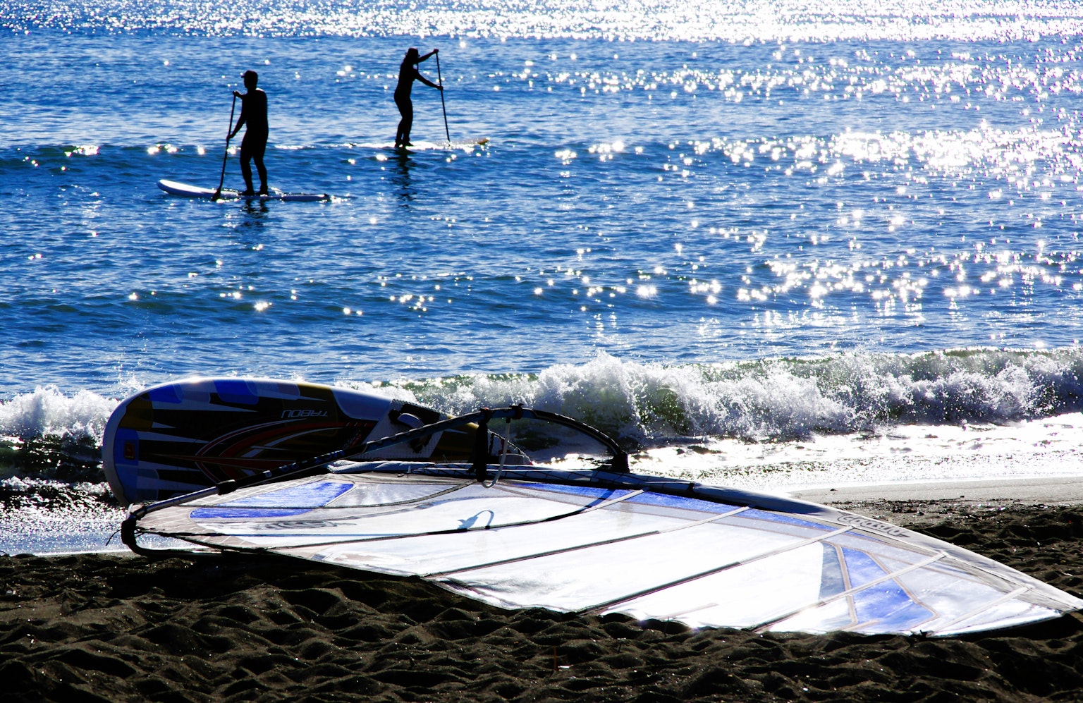 Menschen, die im Ozean Paddelboard fahren, mit einem Windsurfbrett am Sandstrand