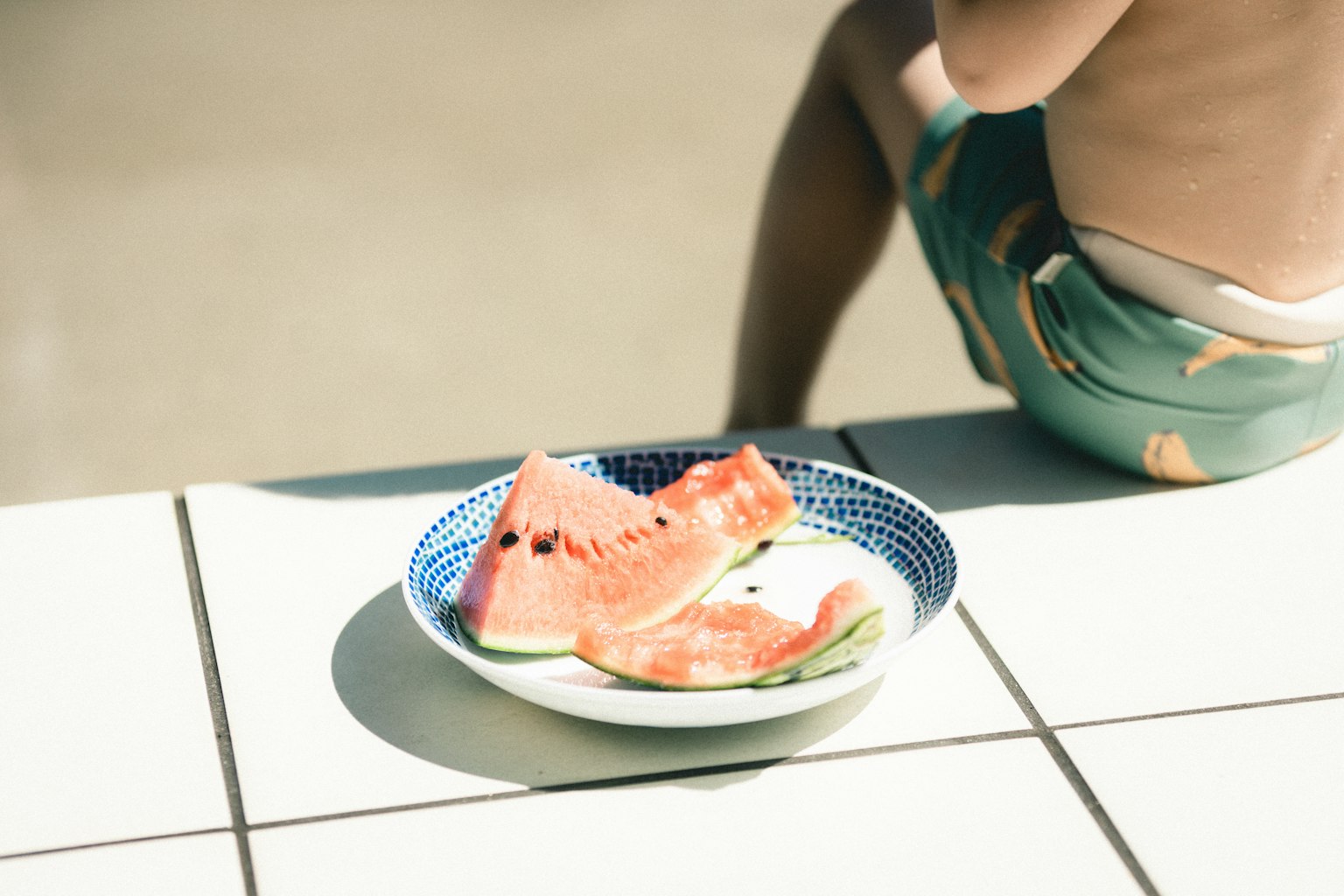 Une assiette de tranches de pastèque au bord de la piscine avec un enfant assis à proximité
