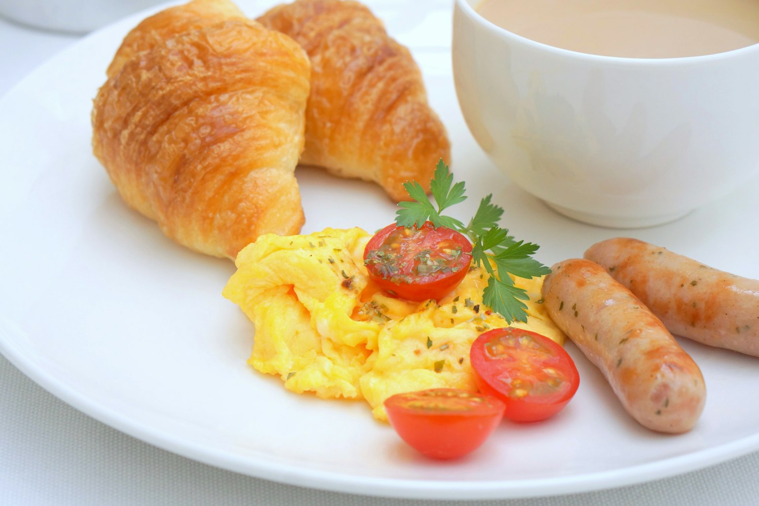Plato de desayuno con croissants salchichas huevos revueltos tomates cherry y una taza de café