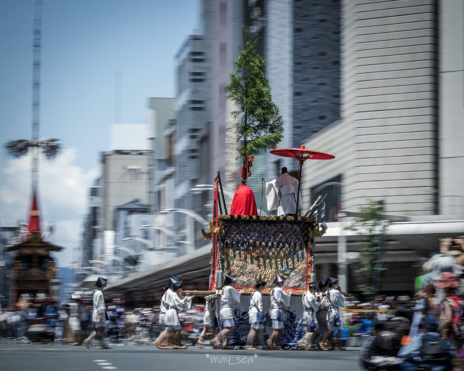 Gambar kereta festival bergerak di jalan dengan seseorang yang memegang payung merah dan pohon hijau
