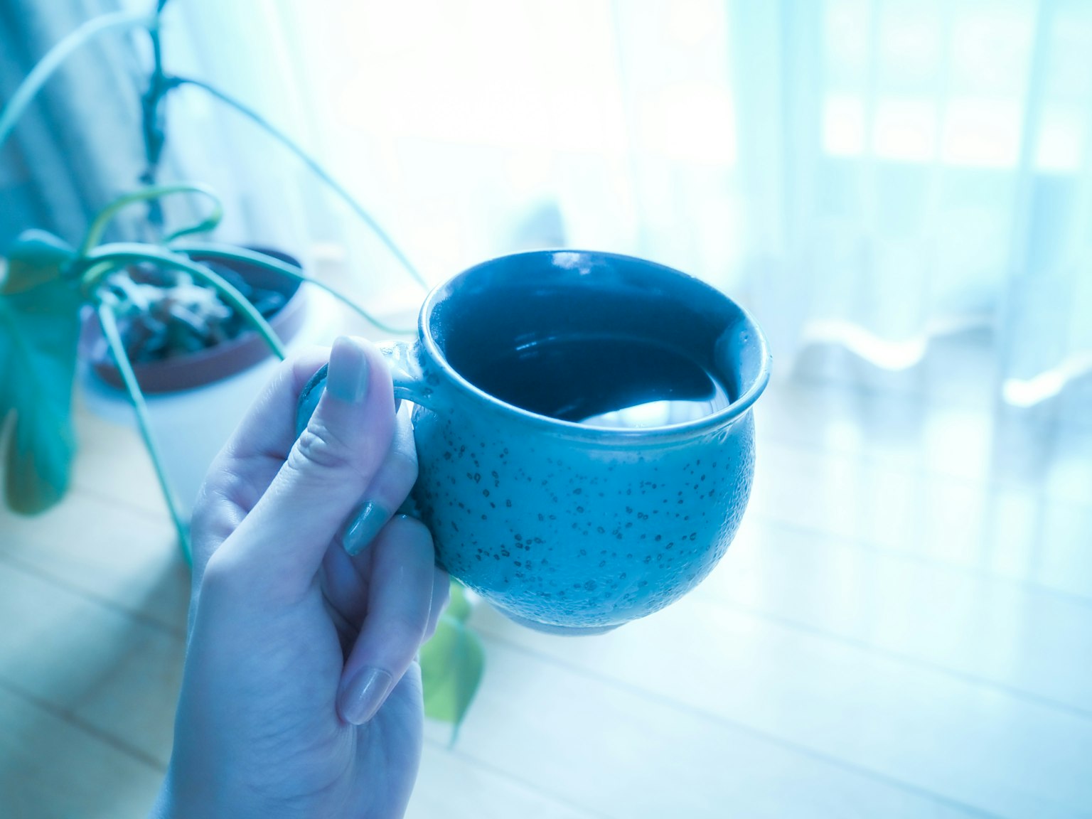 Close-up of a hand holding a blue ceramic cup near a sunlit window