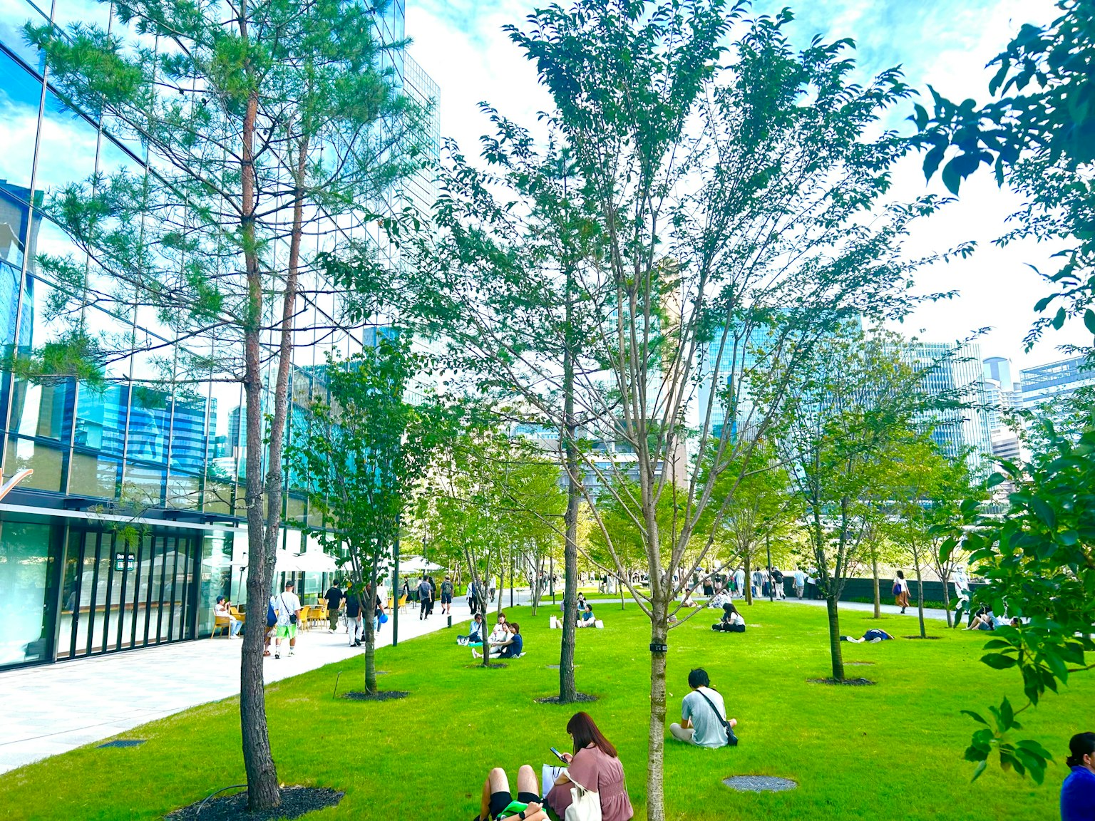 Parco verdeggiante di fronte a edifici moderni con persone sedute tra gli alberi