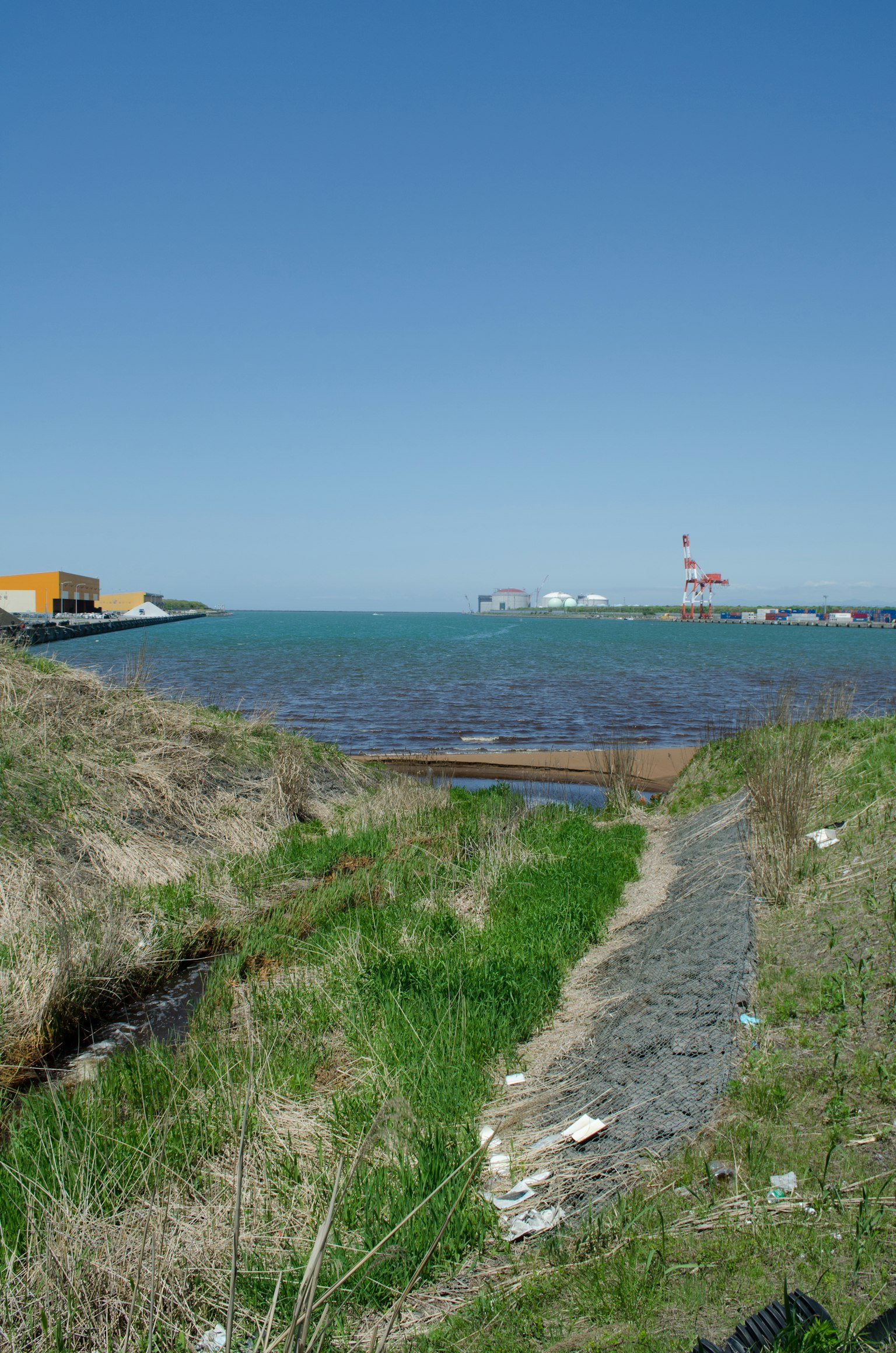 Paysage avec mer bleue et herbe verte, bâtiments portuaires et cheminée rouge en arrière-plan