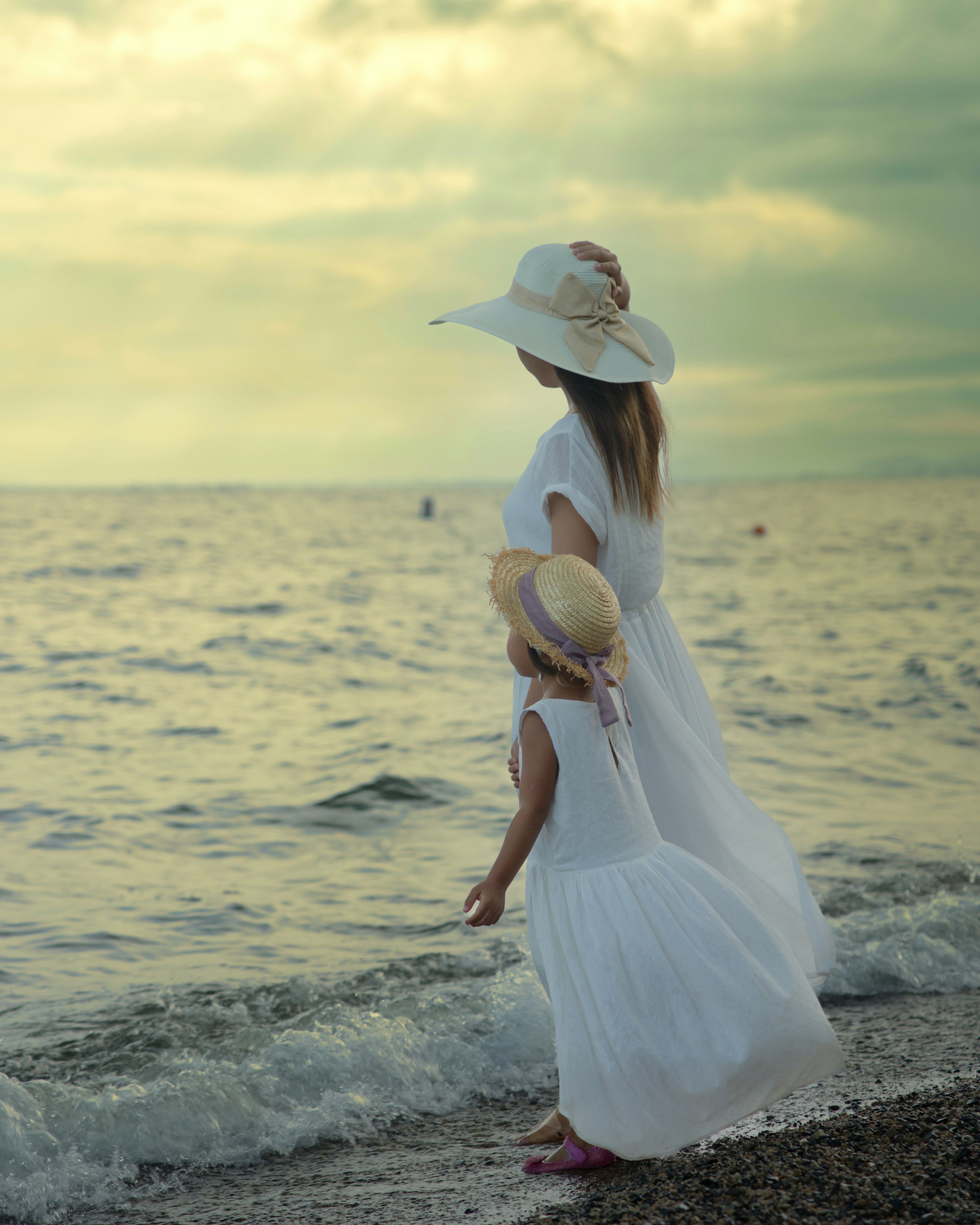 Mother and child in white dresses wearing straw hats by the sea