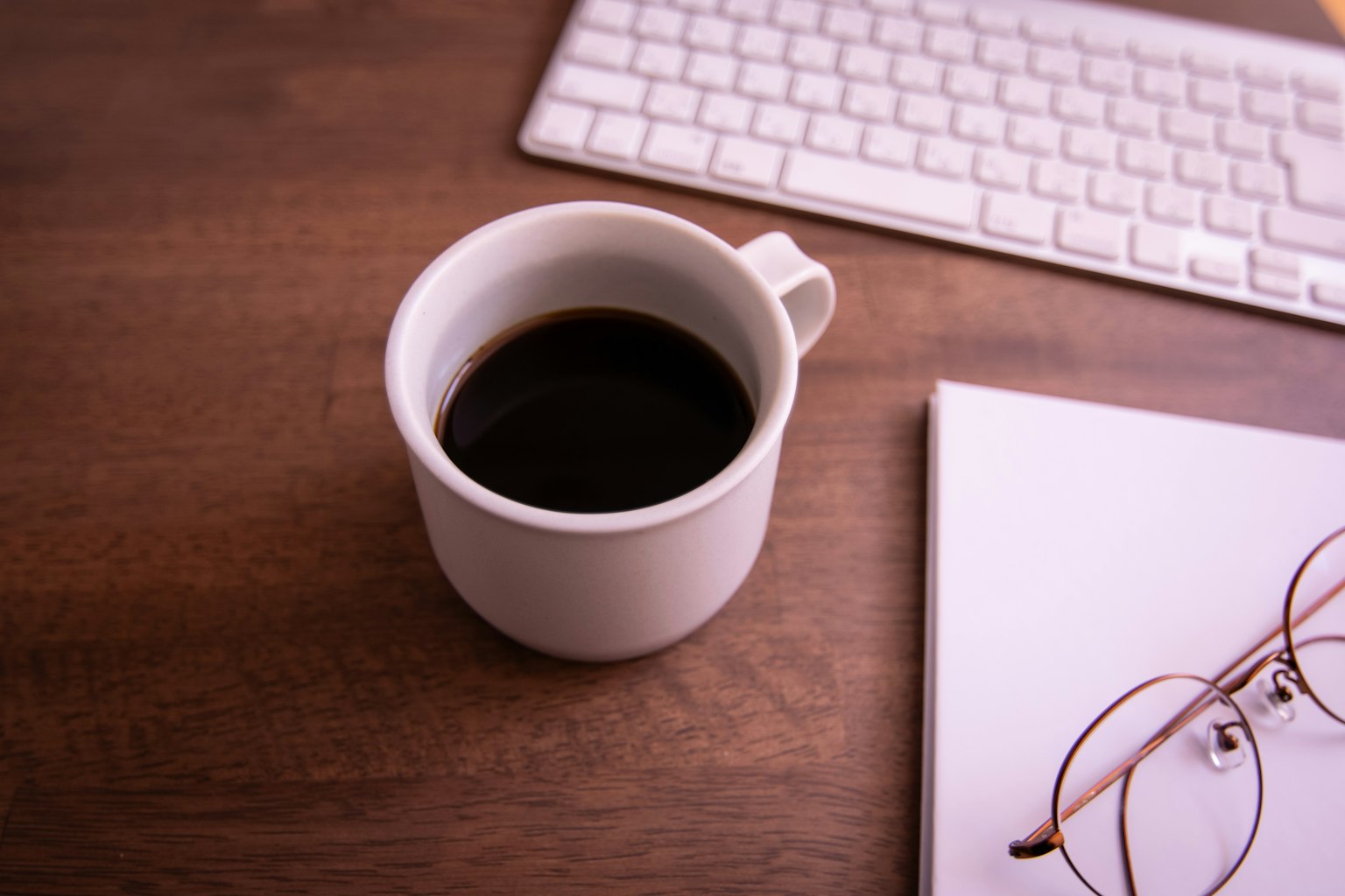 Una tazza bianca di caffè con caffè nero su una scrivania in legno con una tastiera e un quaderno con occhiali