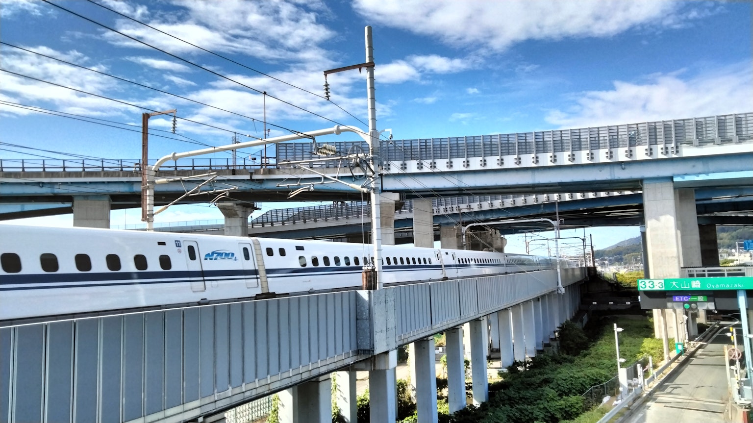 Shinkansen cruzando bajo una carretera elevada con cielo azul