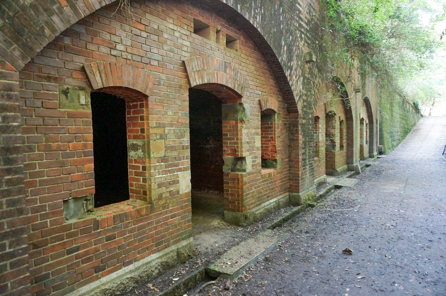 Vista laterale di un vecchio edificio in mattoni con finestre ad arco e vegetazione lussureggiante