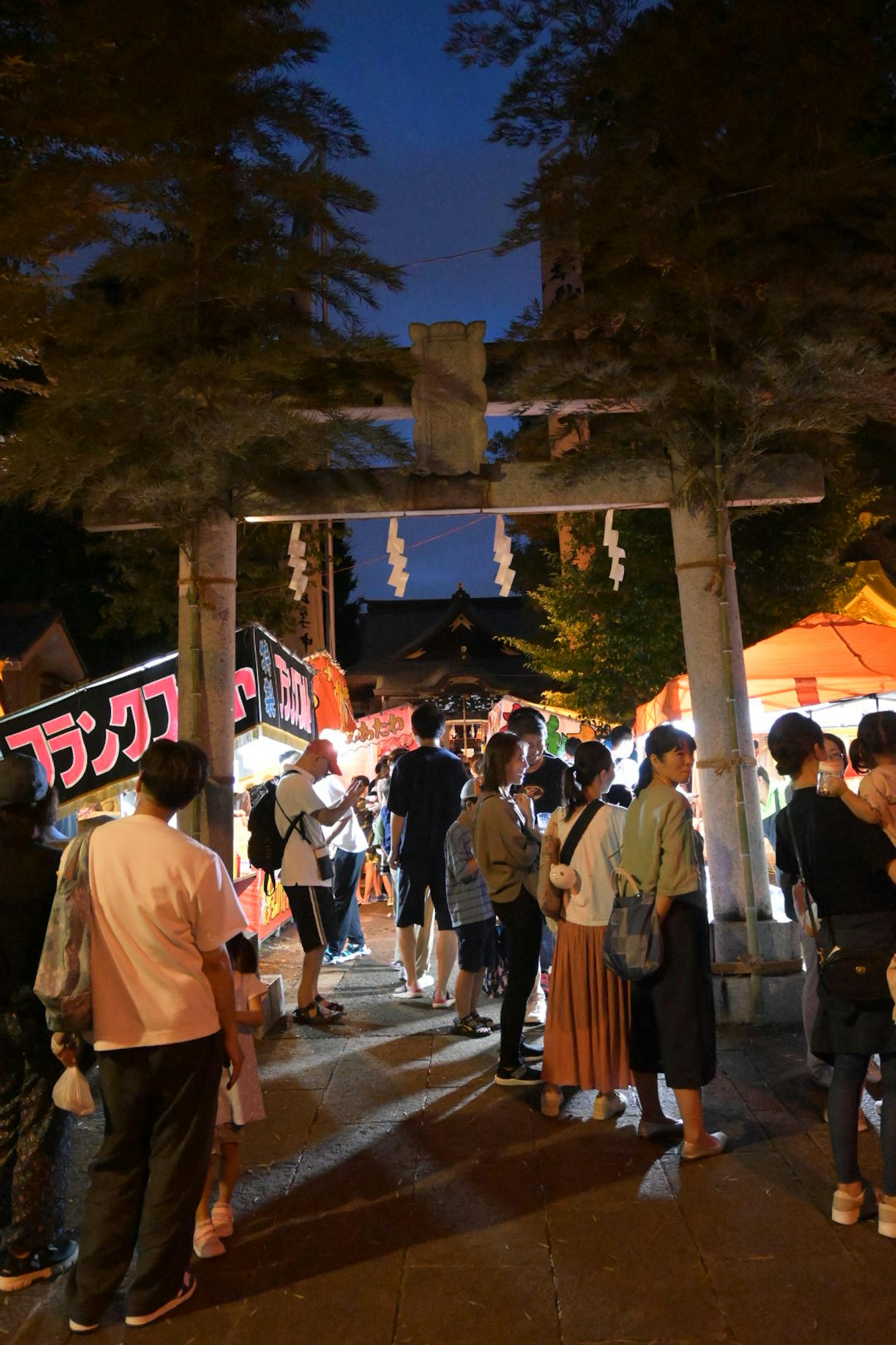 Scène de festival nocturne avec des gens rassemblés autour de stands de nourriture vibrants et d'une porte torii