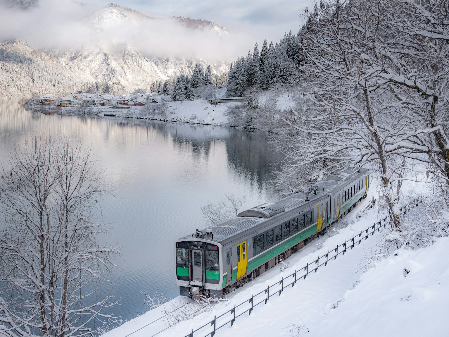 Treno verde e giallo che corre lungo un lago innevato e montagne
