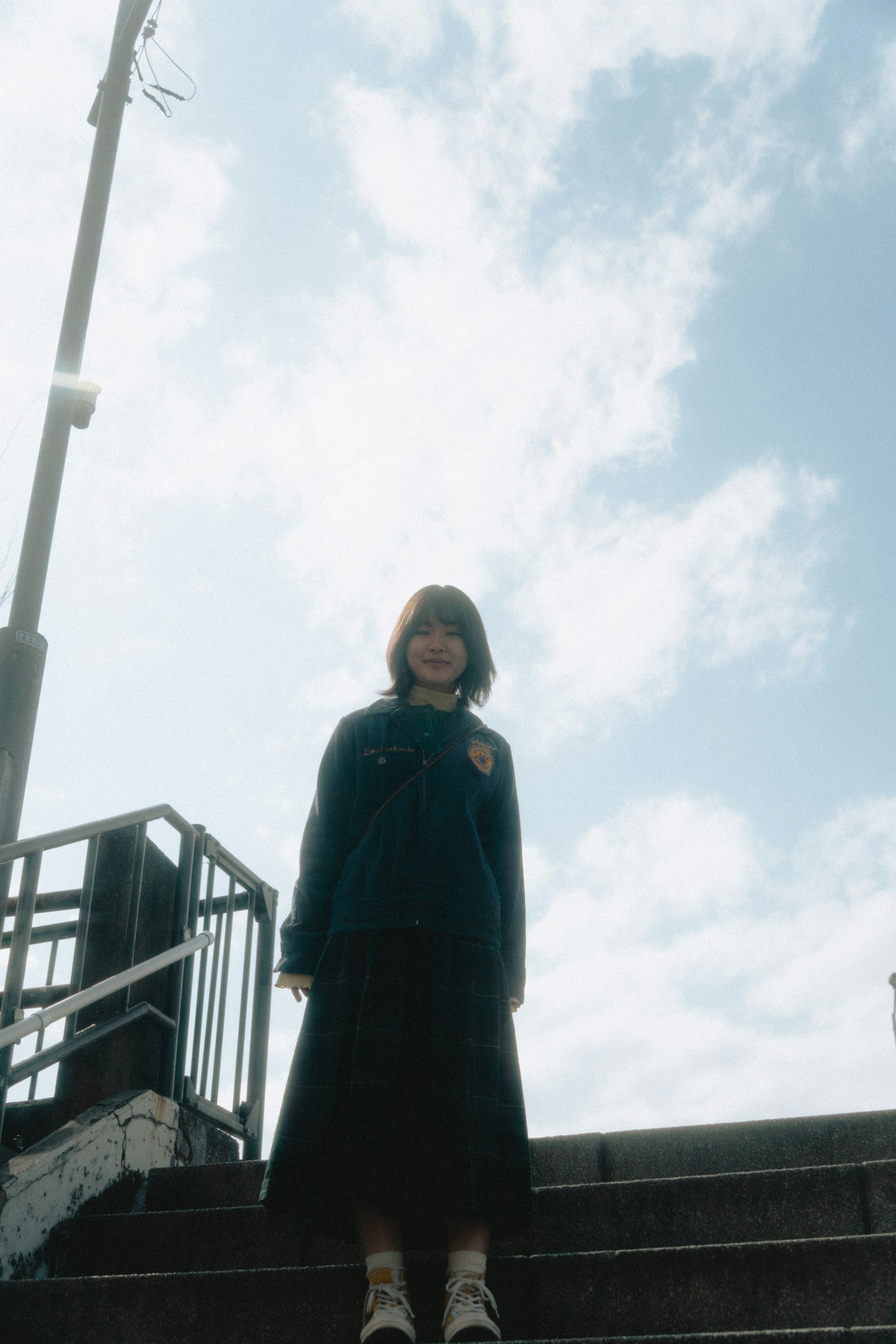 Eine Frau in blauer Jacke steht auf einer Treppe vor einem Hintergrund aus blauem Himmel und weißen Wolken