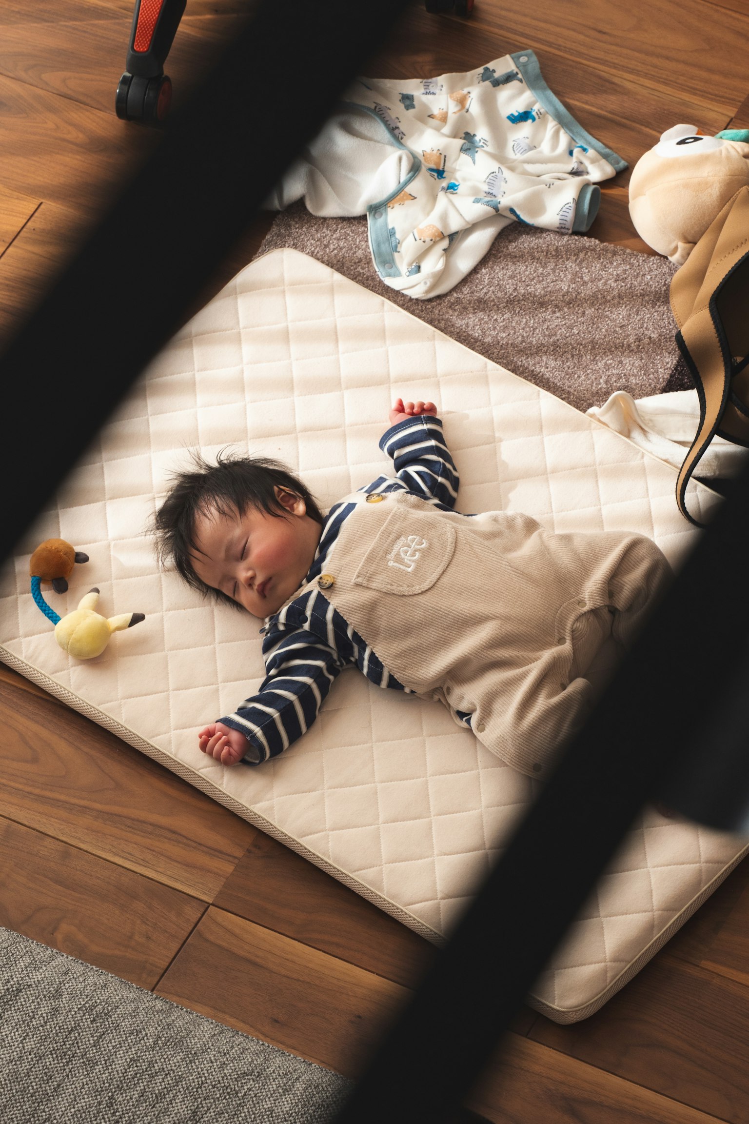 Seorang bayi tidur di atas matras mengenakan baju garis-garis dan overall dengan mainan di dekatnya