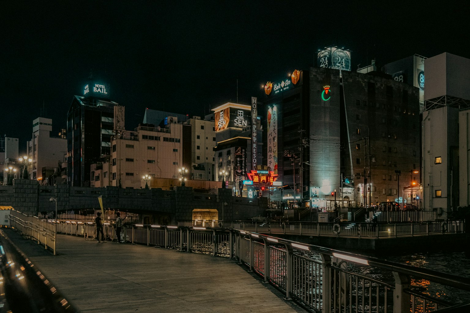 Nighttime cityscape along a riverside walkway with buildings