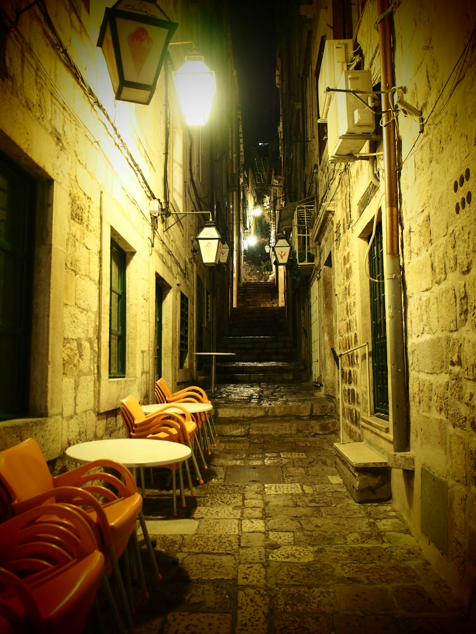Callejón estrecho de noche con sillas y mesas apiladas Paredes de piedra y farolas vintage crean una atmósfera encantadora