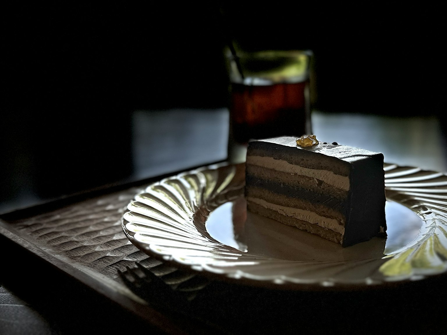 Ein Stück Schokoladenkuchen auf einem kunstvoll verzierten Teller mit einem Getränk in einer schwach beleuchteten Umgebung
