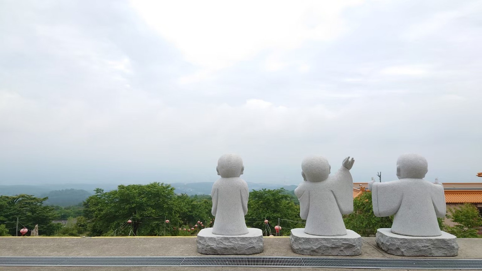 Drei weiße Buddha-Statuen sitzen mit Blick auf grüne Bäume und bewölkten Himmel